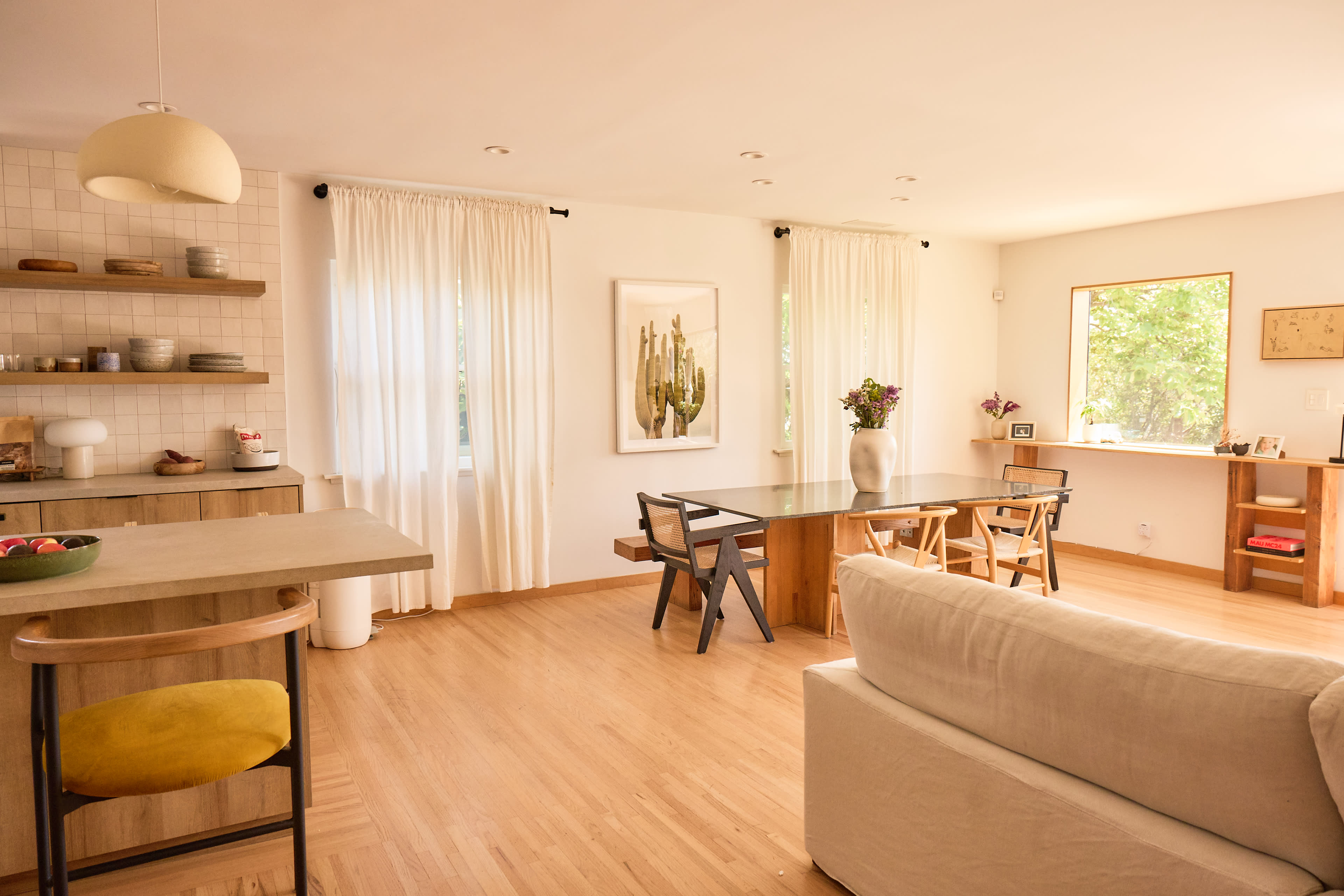 The image shows a bright, modern living space featuring a dining table, chairs, and a kitchen area, all adorned with large windows and minimalist decor.