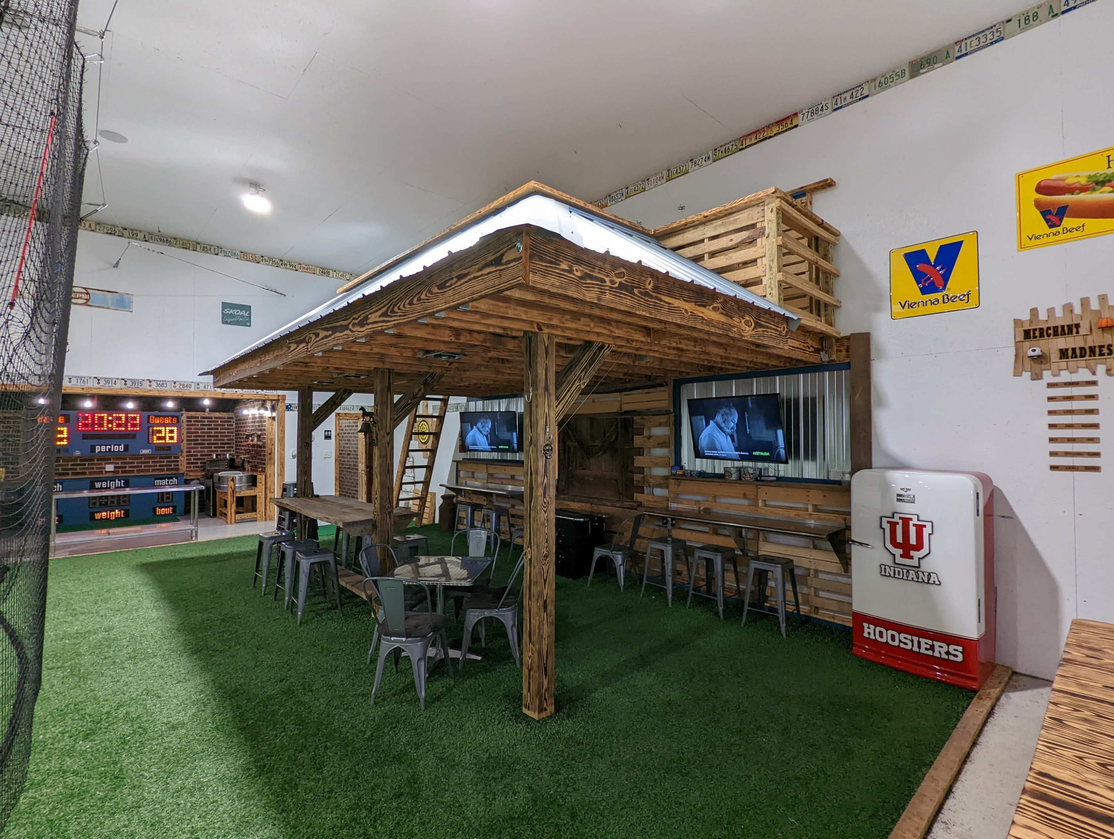 The image shows an indoor recreation area featuring a wooden structure with a roof, seating, and two large televisions, surrounded by artificial grass and decorated with various sports-themed items.