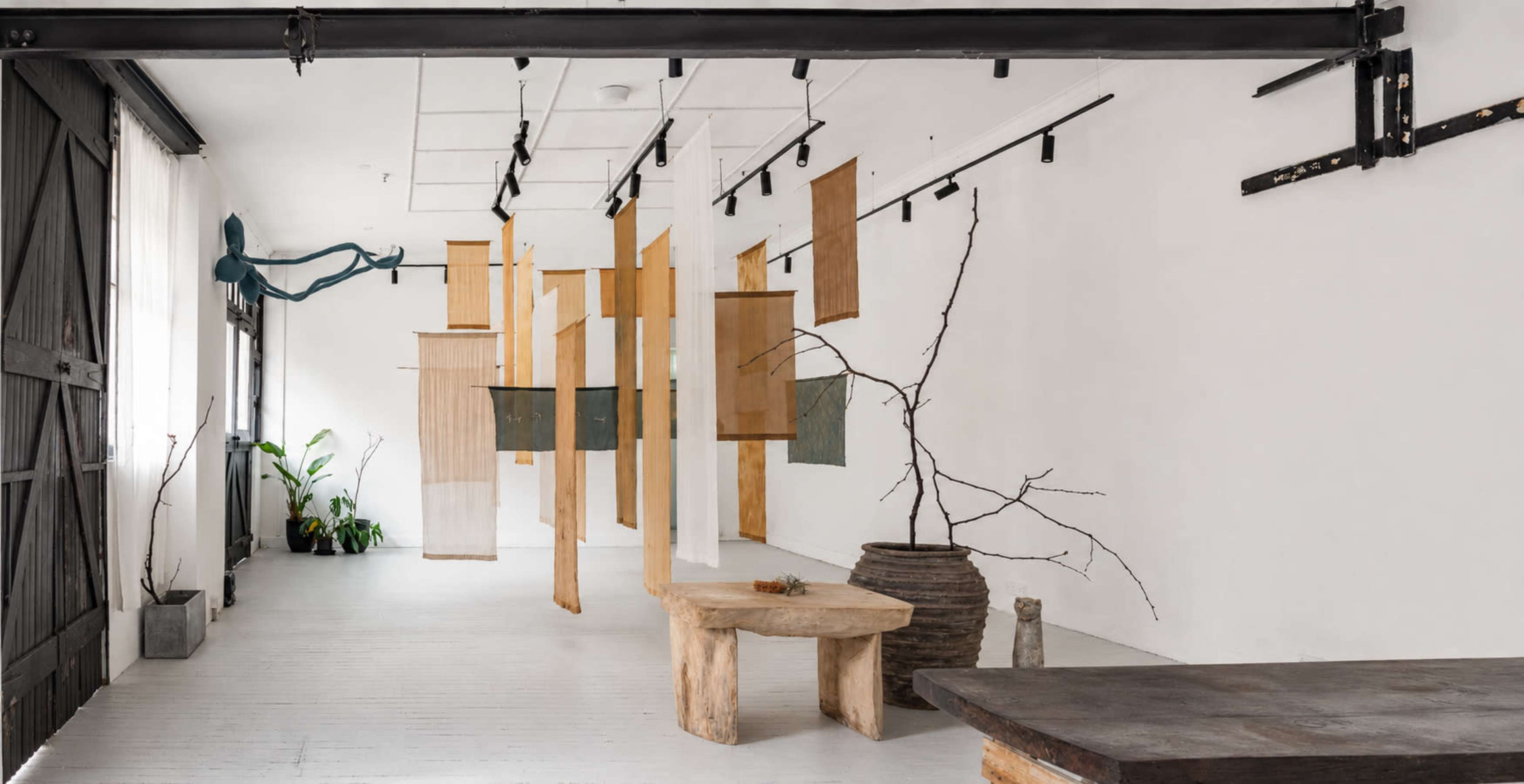 The image shows a minimalist art gallery with hanging abstract panels, natural wood furniture, and decorative elements like branches and potted plants.