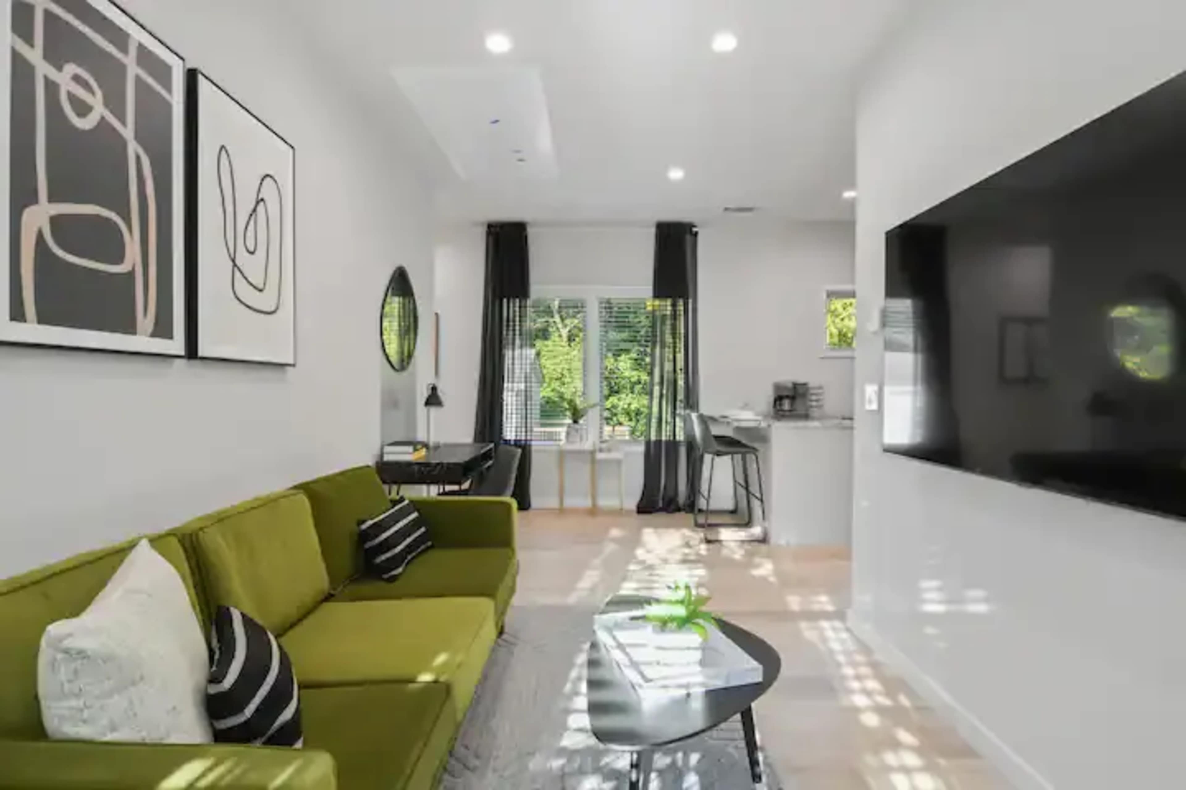 A modern living room features a green sofa, abstract wall art, and a large flat-screen TV, with sunlight streaming through the windows.