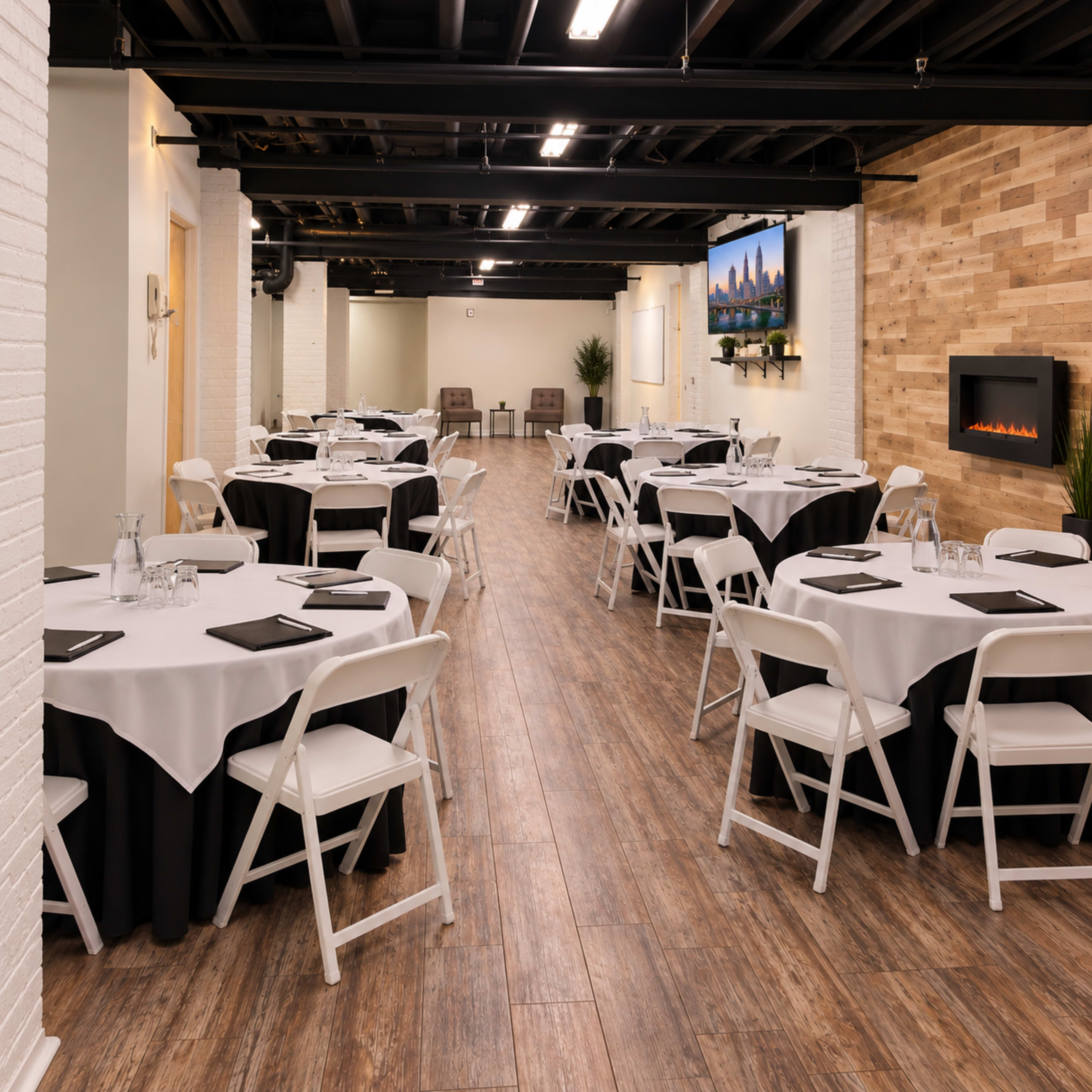 A spacious meeting room features round tables dressed in white tablecloths with black skirts, set up in rows along a wooden floor, with a fireplace and artwork on the walls.