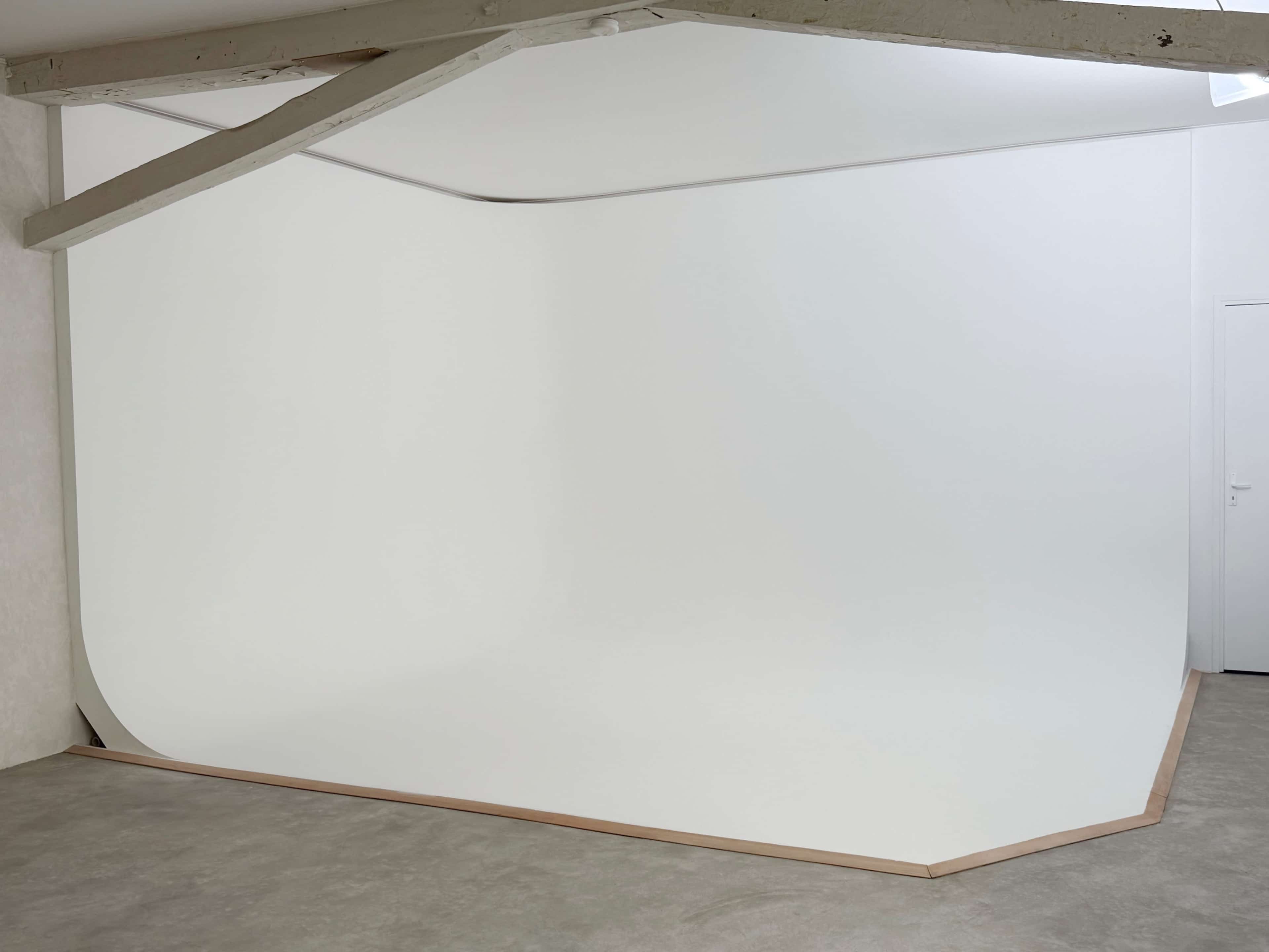 The image shows a minimalistic studio space featuring a seamless white backdrop that curves into the floor, complemented by wooden trim along its edges.