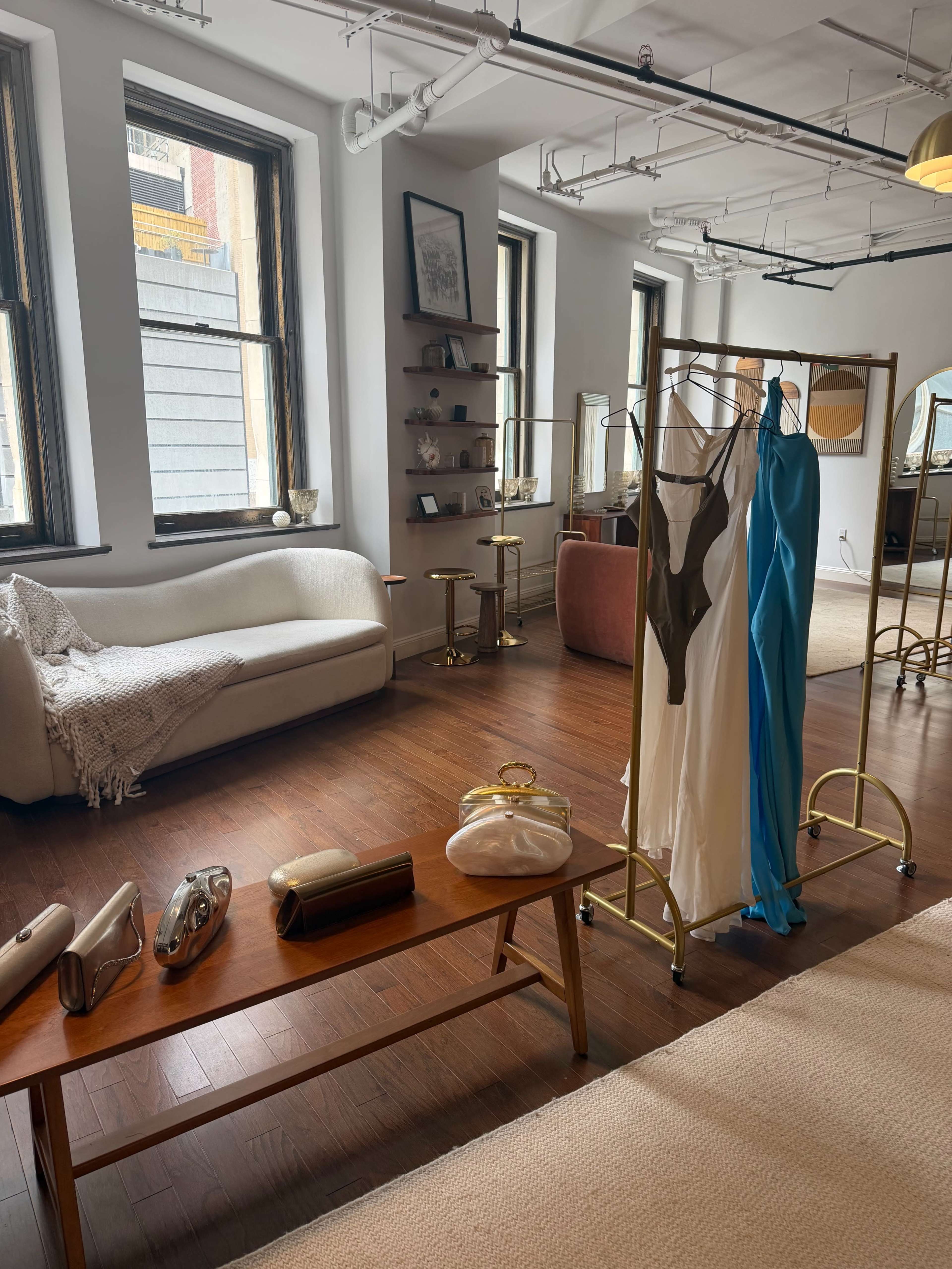 The image shows a spacious, well-lit room featuring a couch, a wooden table with various bags, and a clothing rack displaying several dresses.