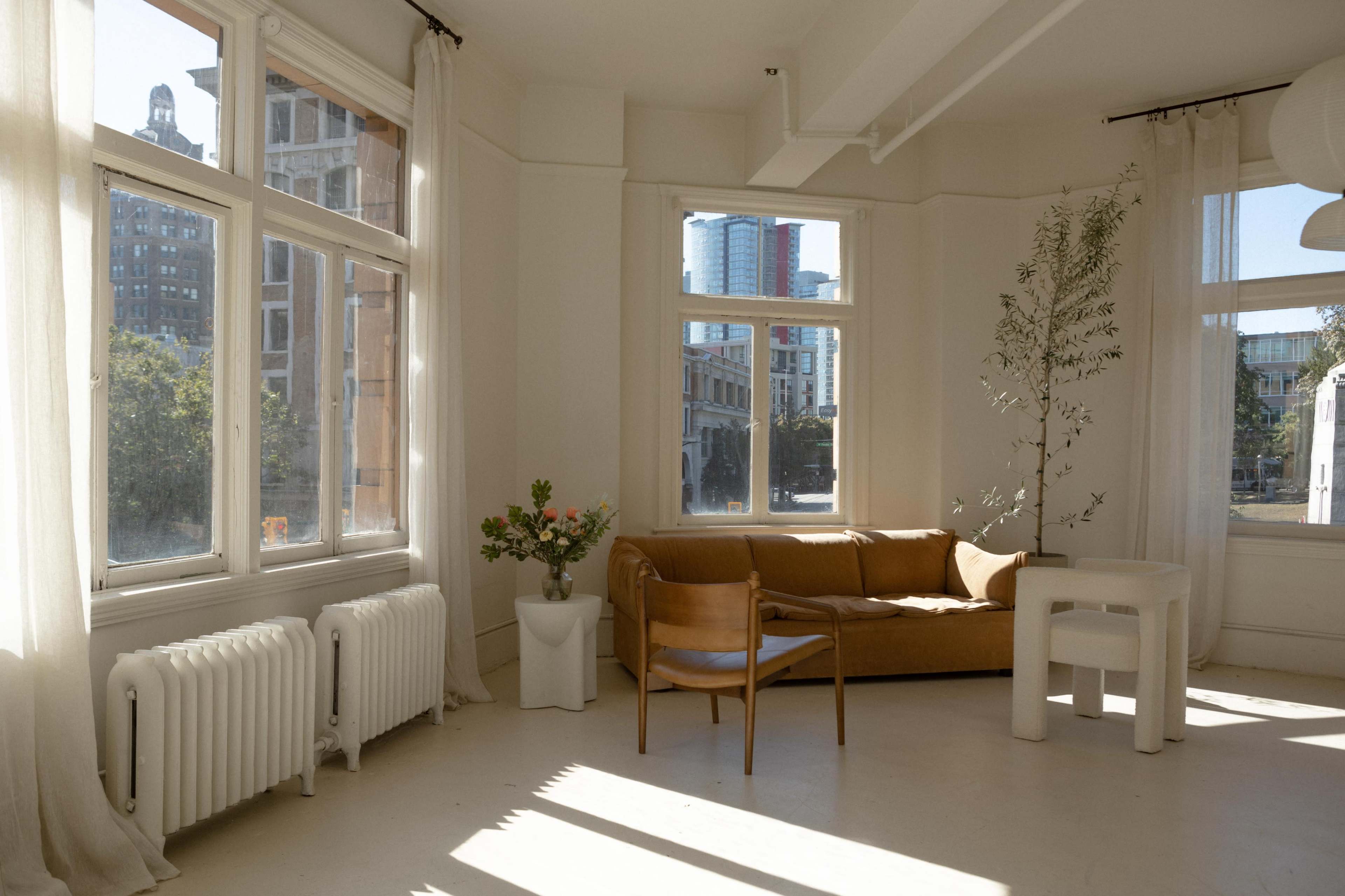 A bright, minimalist interior features a brown couch, a wooden chair, and a white table beside large windows with soft curtains.