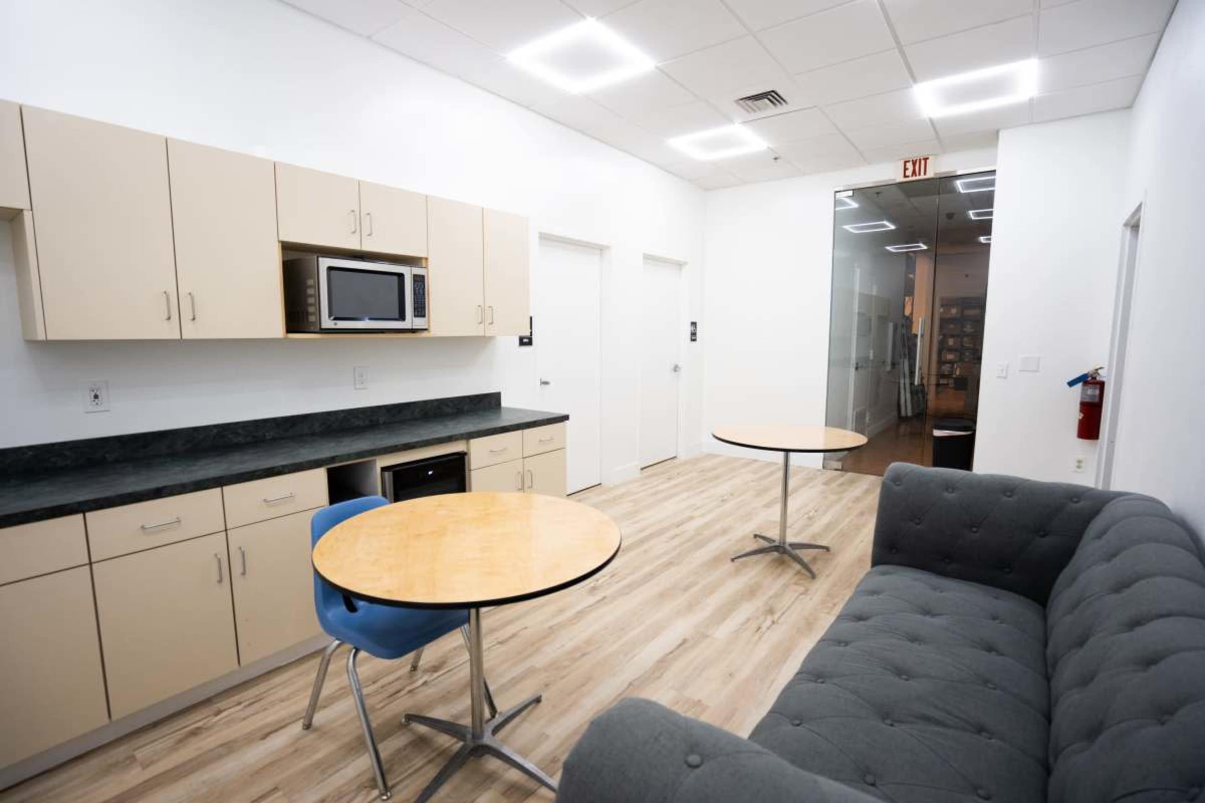 The image depicts a modern kitchenette area featuring a microwave, a refrigerator, two round wooden tables, and a gray couch in an open layout.