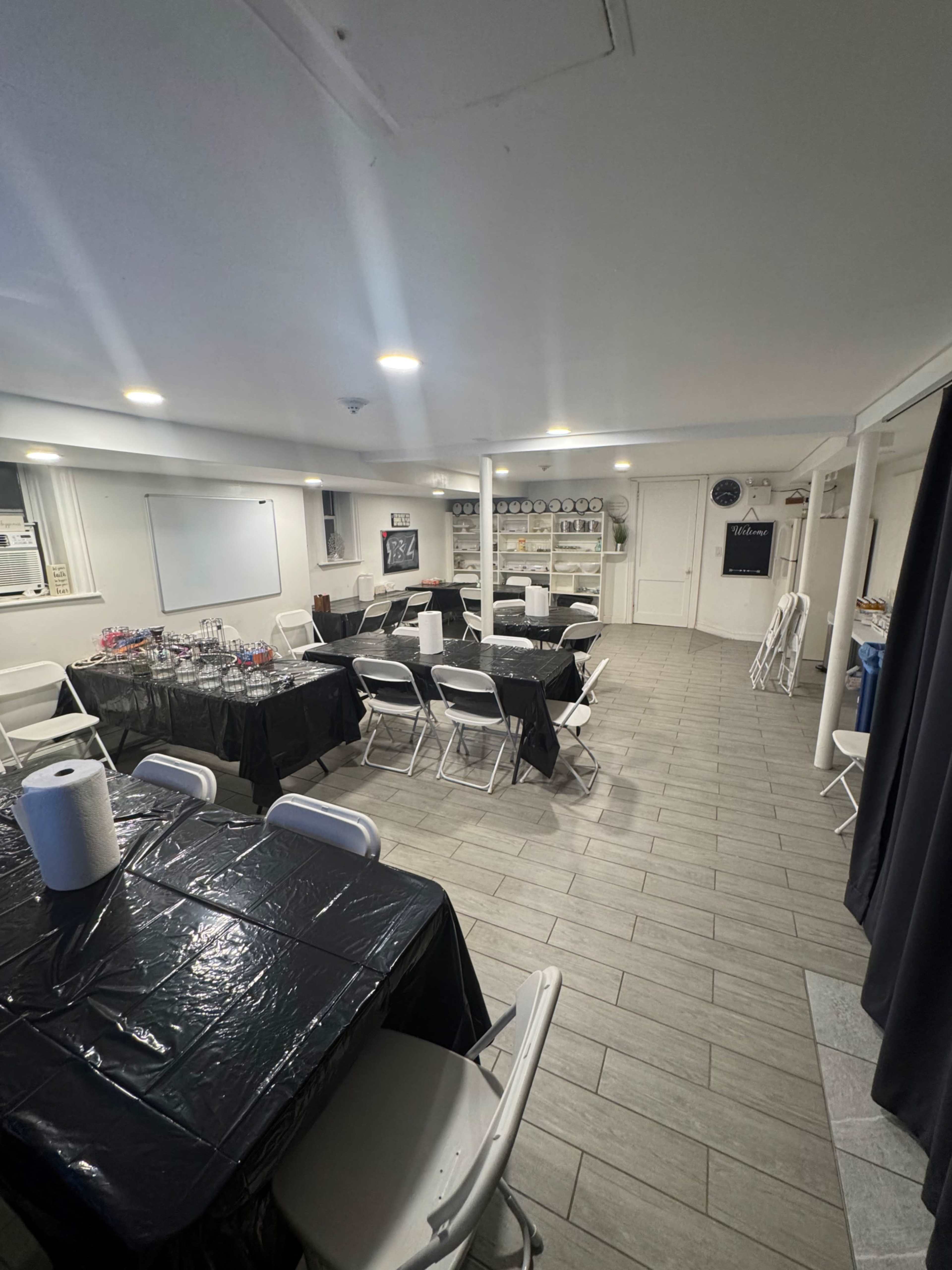 The image shows a spacious room with several tables covered in black tablecloths and arranged with white chairs, alongside a whiteboard and various items on shelves.