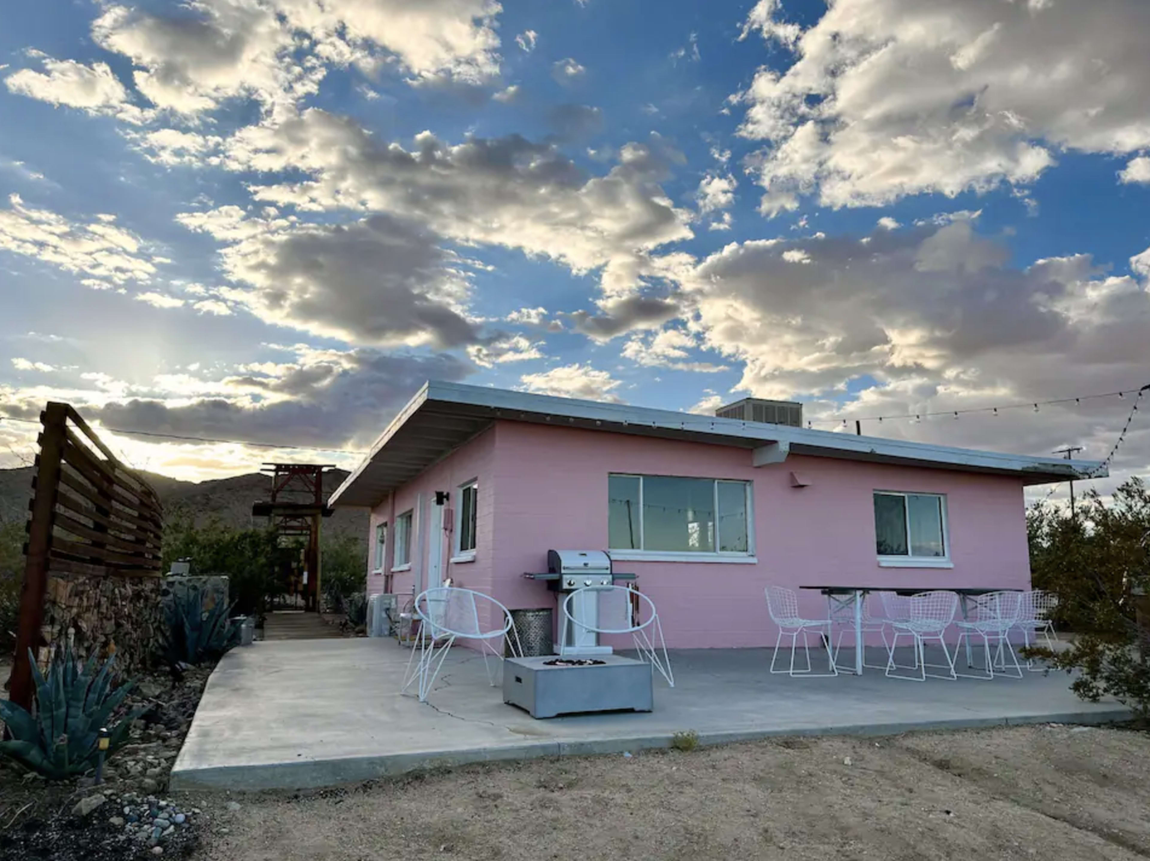 A pink house with a grill and outdoor seating is set against a backdrop of cloudy skies and mountains.