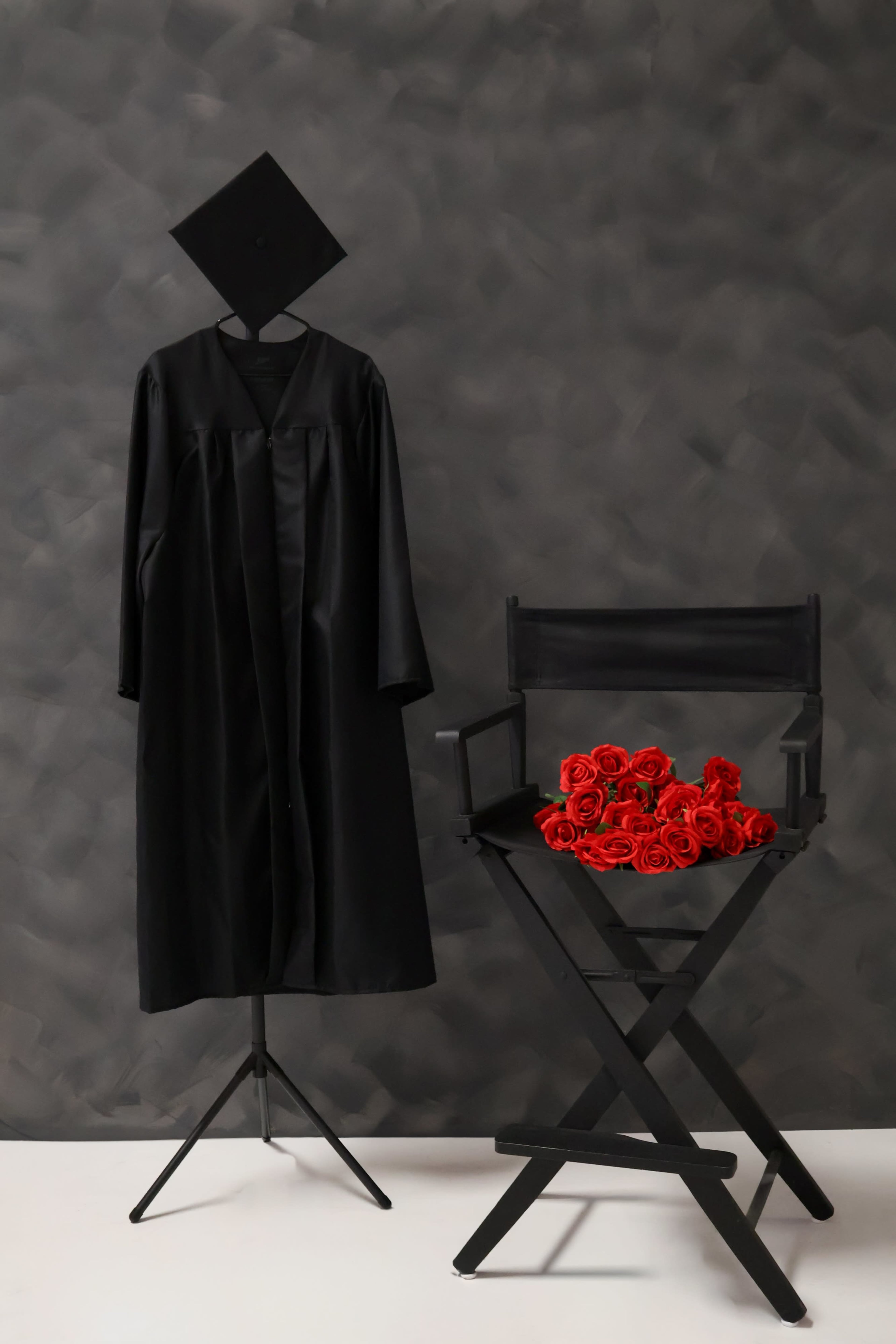 A black graduation gown and cap are displayed on a stand next to a director's chair adorned with a bouquet of red roses.