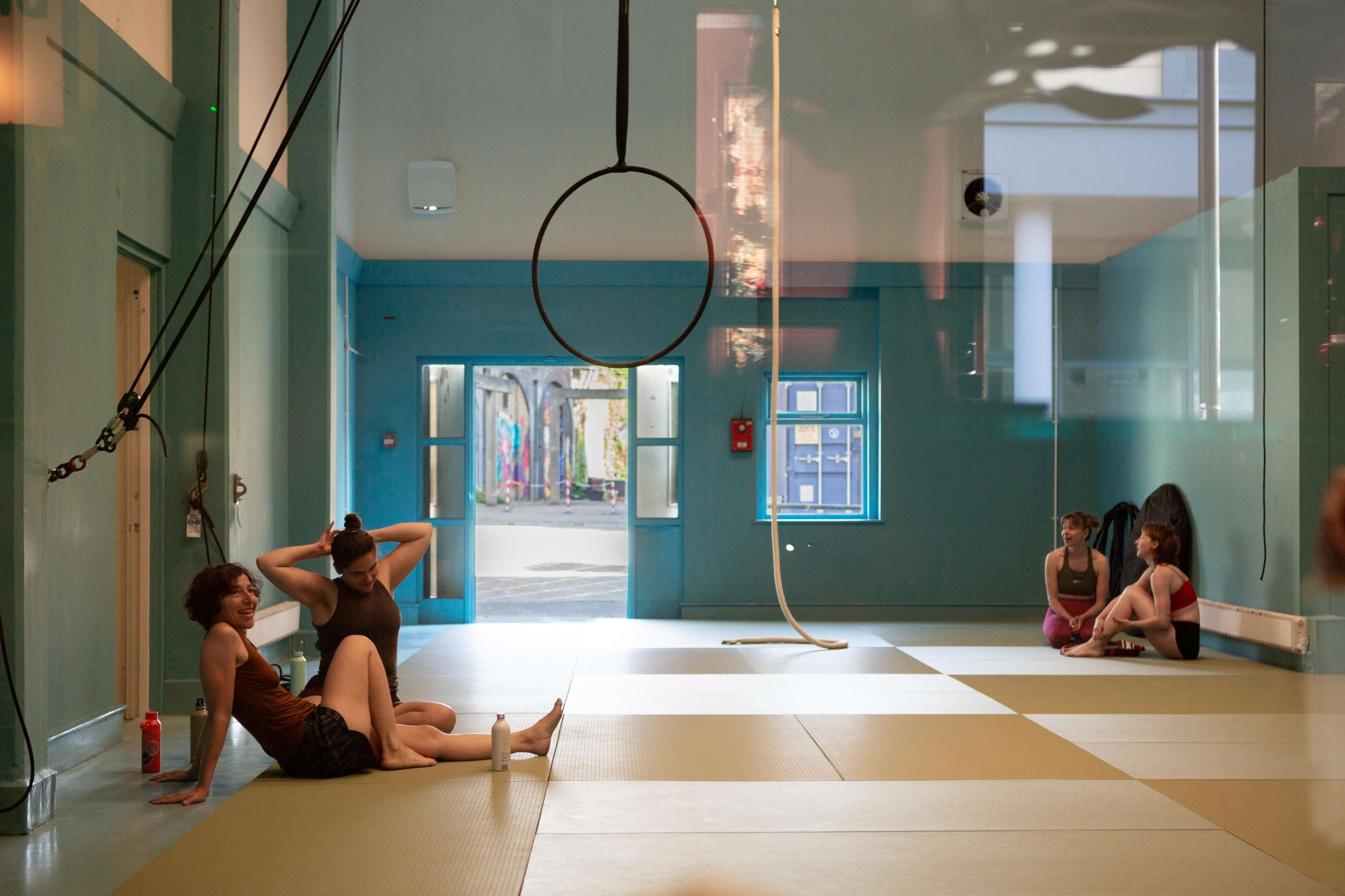 The image shows a spacious indoor gym or studio with a light blue color scheme, featuring several people engaged in leisure activities and aerial apparatus hanging from the ceiling.