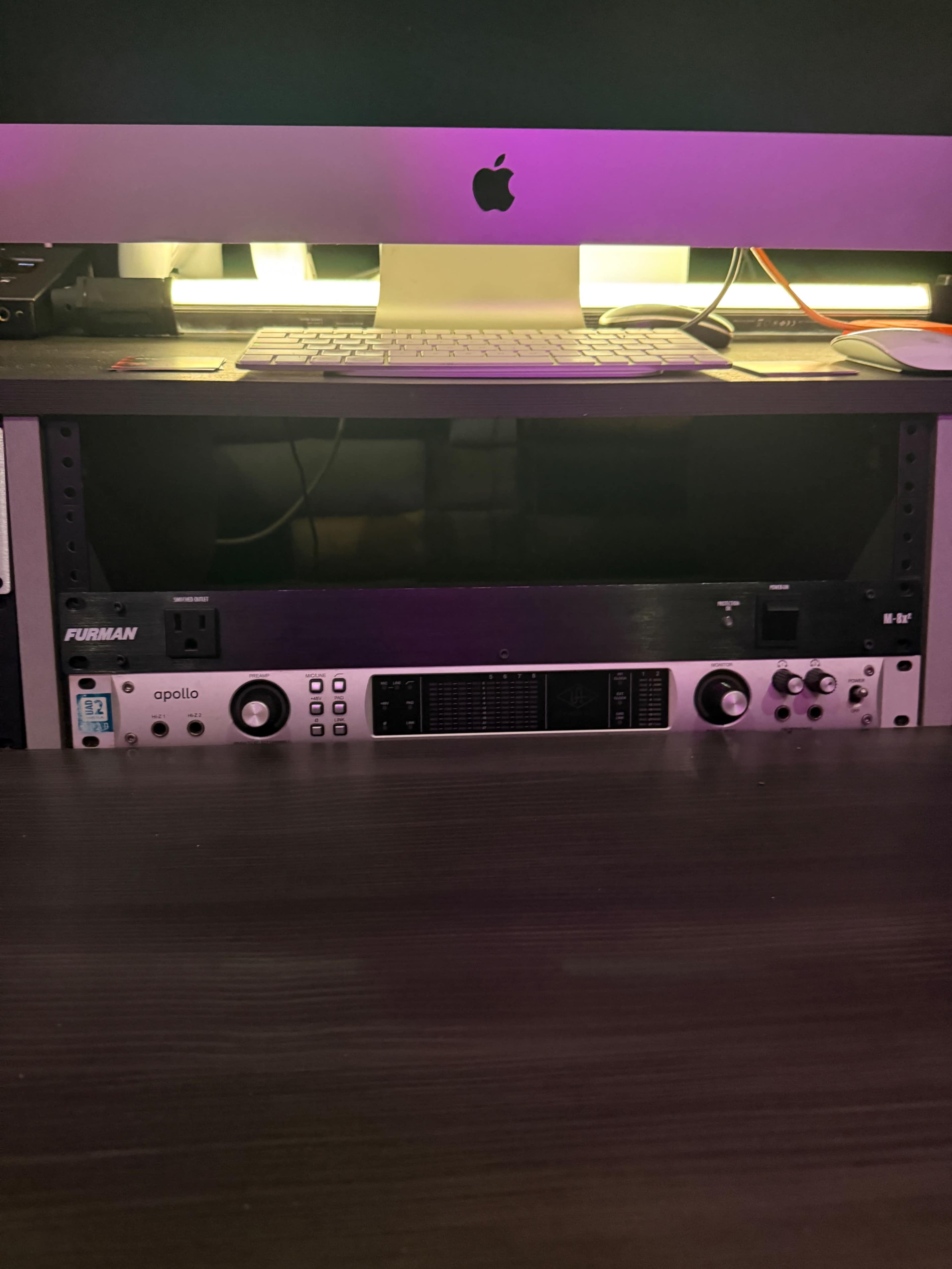 The image shows a desktop setup featuring an Apple computer and an audio interface placed on a shelf below it.