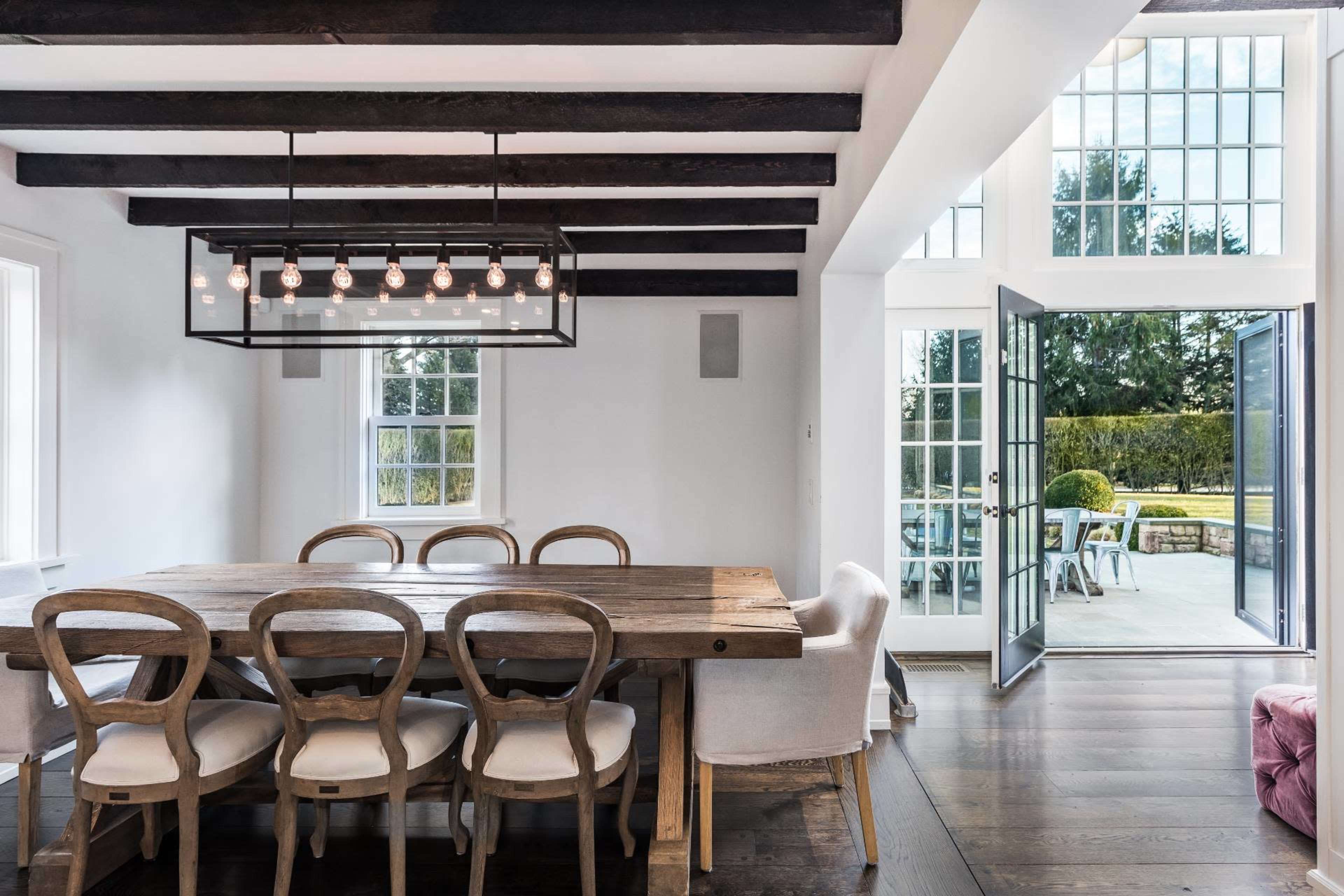 A modern dining area features a wooden table surrounded by six chairs, with a pendant light overhead and open doors leading to an outdoor patio.