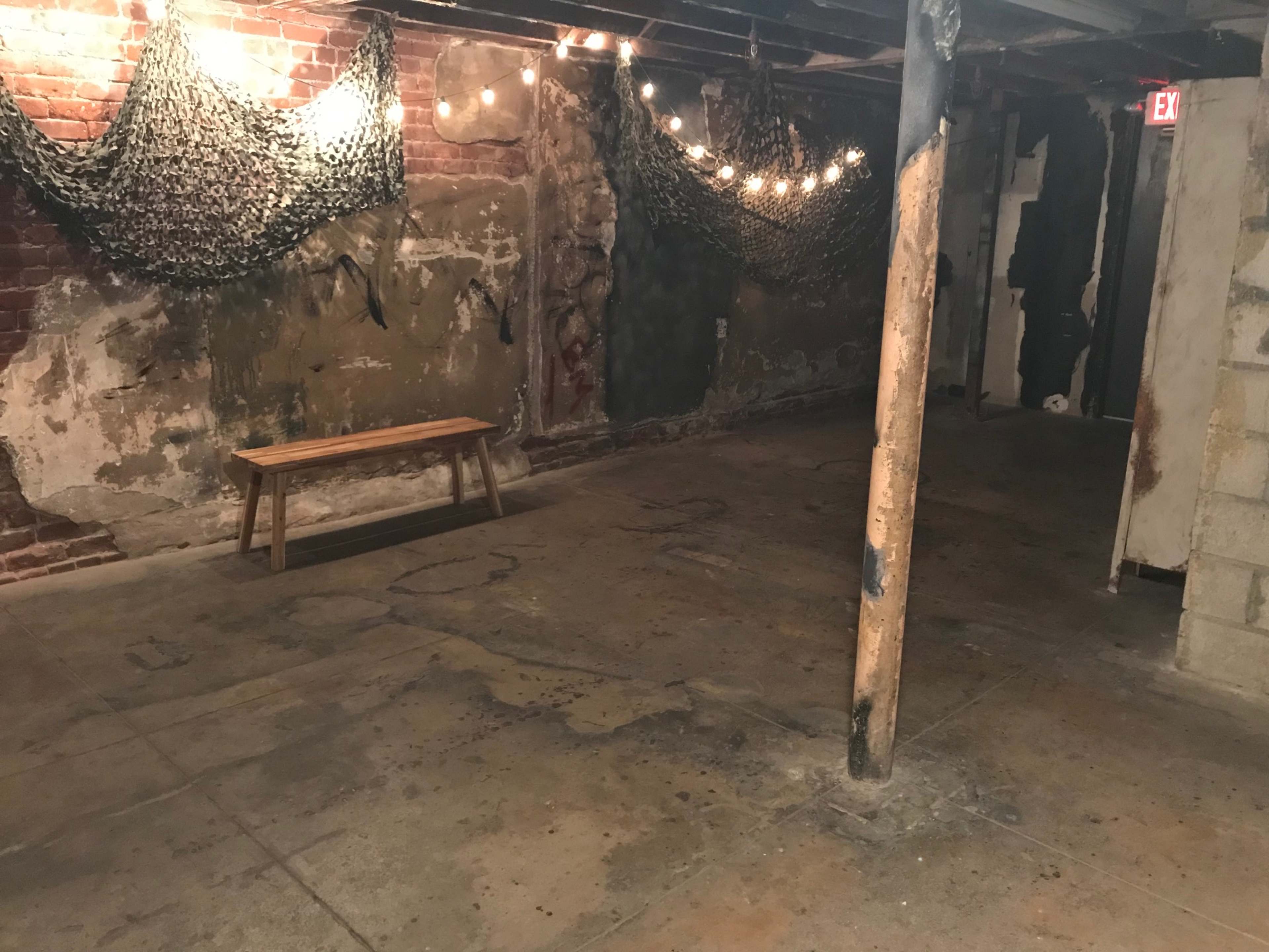 A dimly lit basement with exposed brick walls, a wooden bench, hanging string lights, and a concrete floor.