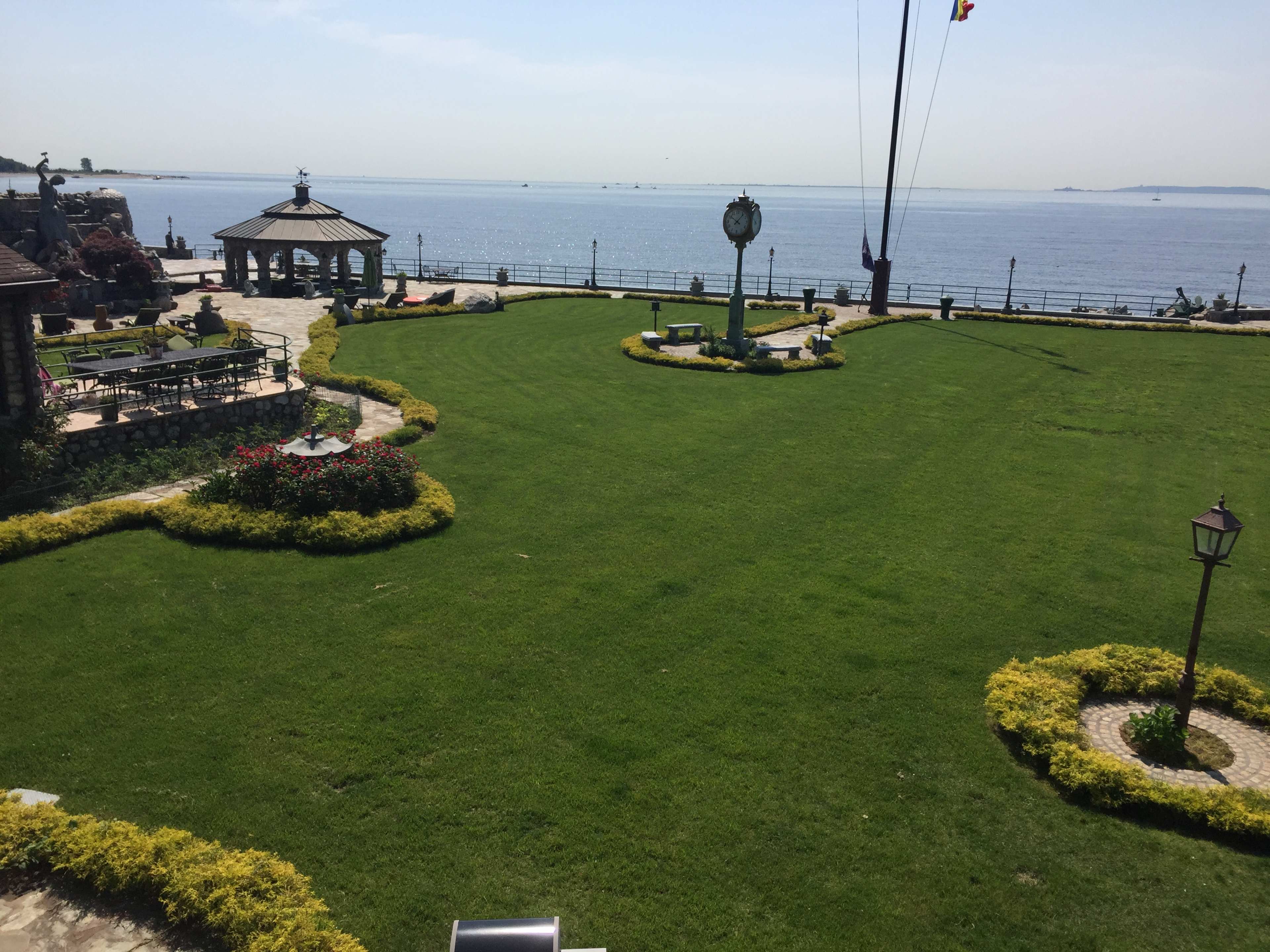 The image shows a well-maintained green lawn with flower beds and a clock tower facing a body of water.