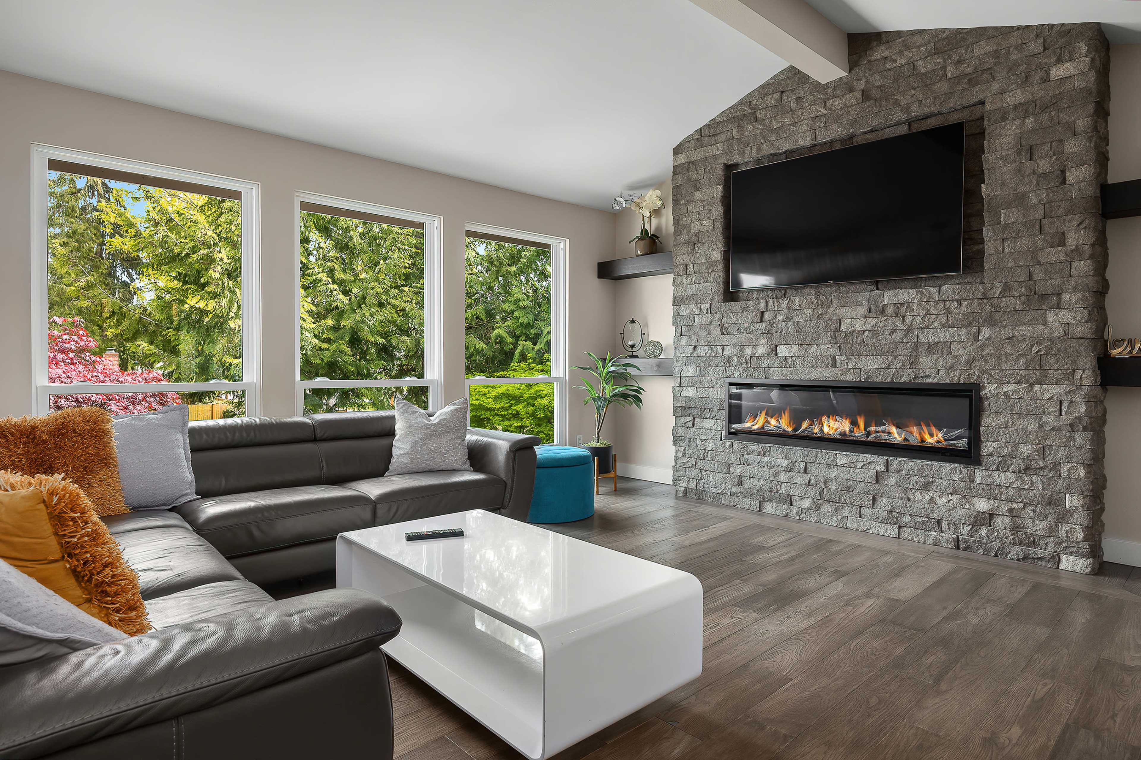 The living room features a modern design with a stone fireplace, a wall-mounted television, large windows, a sectional sofa, and a sleek coffee table.