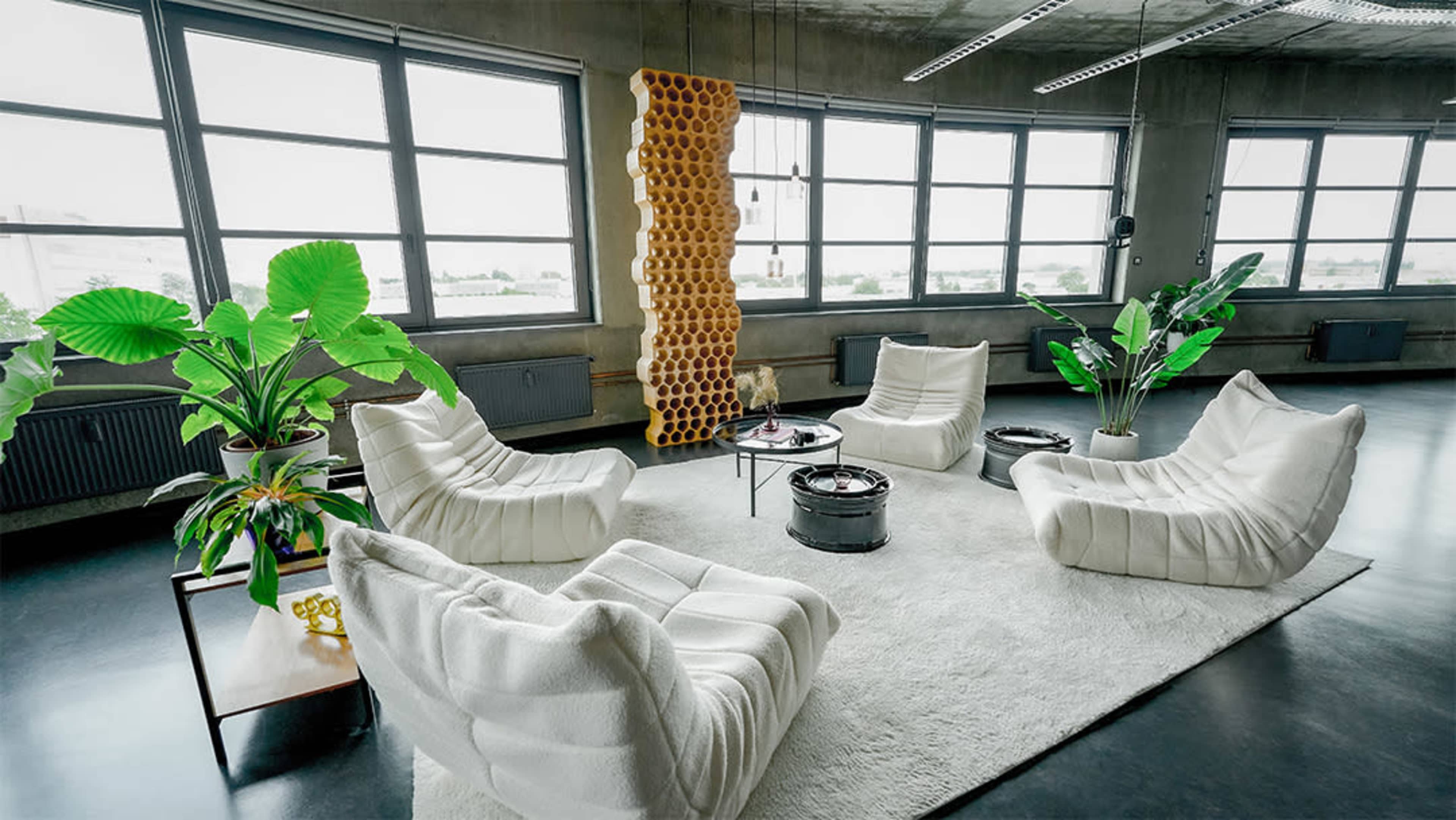 The image displays a modern living space featuring four white, tufted chairs arranged around a circular table on a plush rug, with large windows and potted plants in the background.