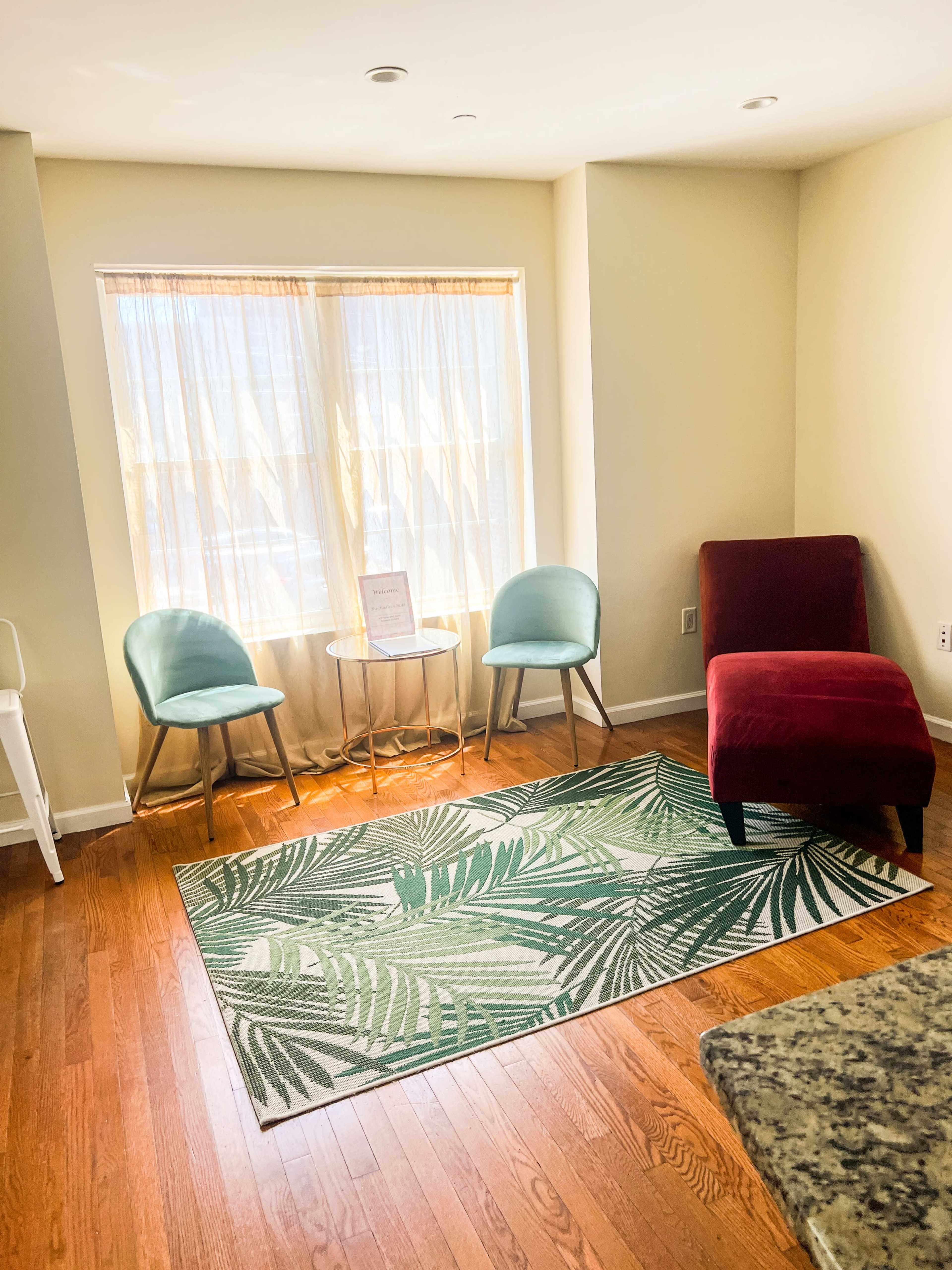 A cozy corner features two chairs and a red chaise lounge near a window with sheer curtains, alongside a small round table on a tropical-patterned rug.