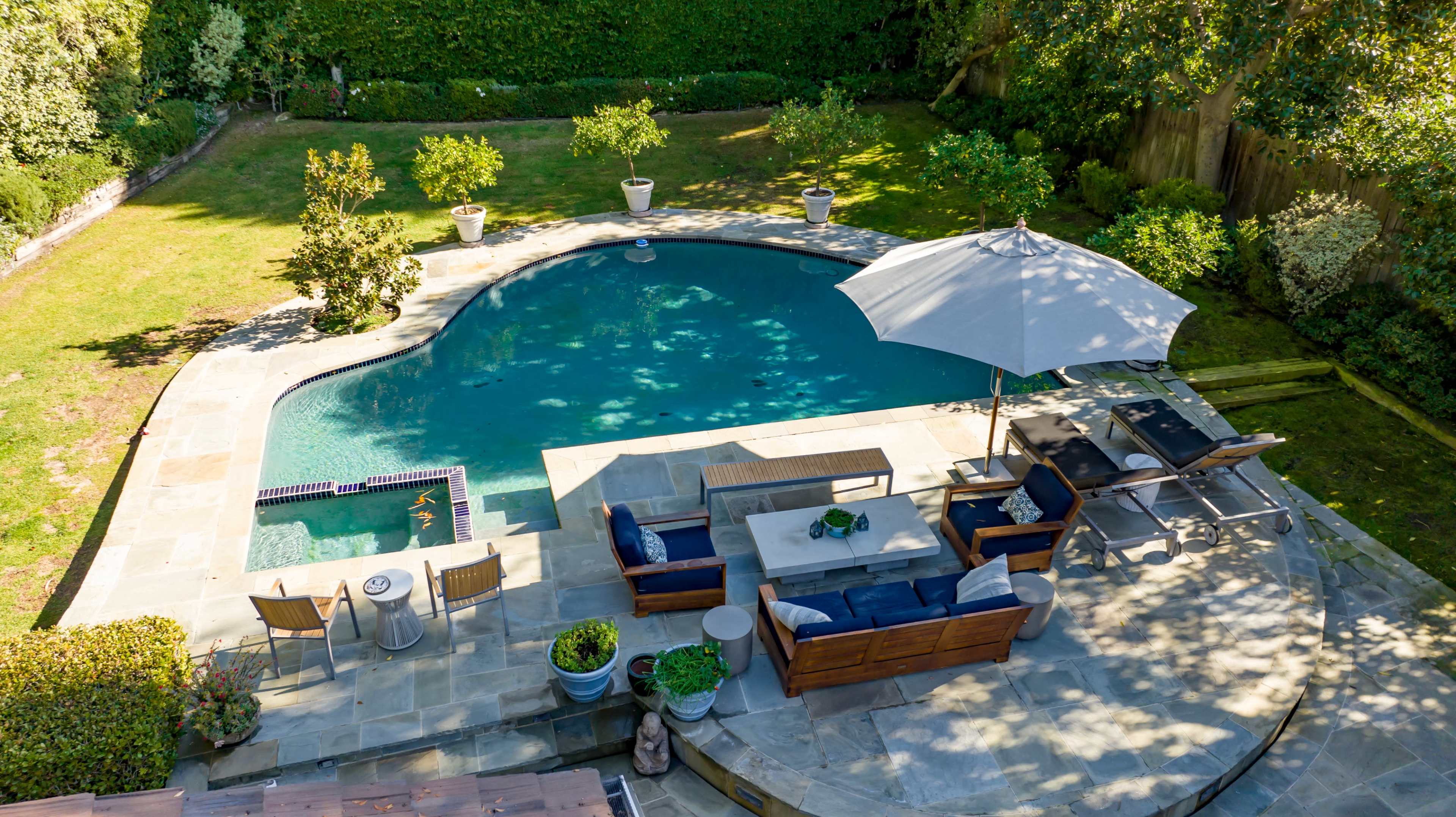 Aerial view of a backyard featuring a curved swimming pool surrounded by a stone patio, lounge furniture, and a garden area with potted plants.