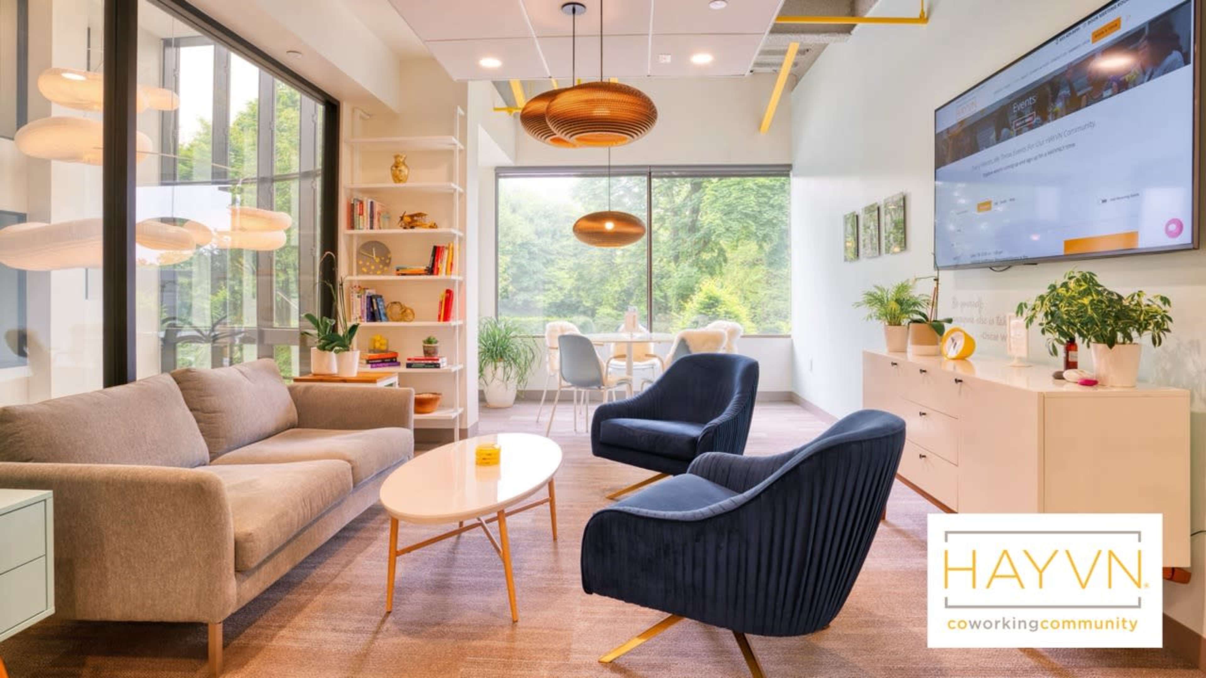 The image shows a modern co-working space with a gray sofa, two dark blue chairs, a white coffee table, and large windows that let in natural light.