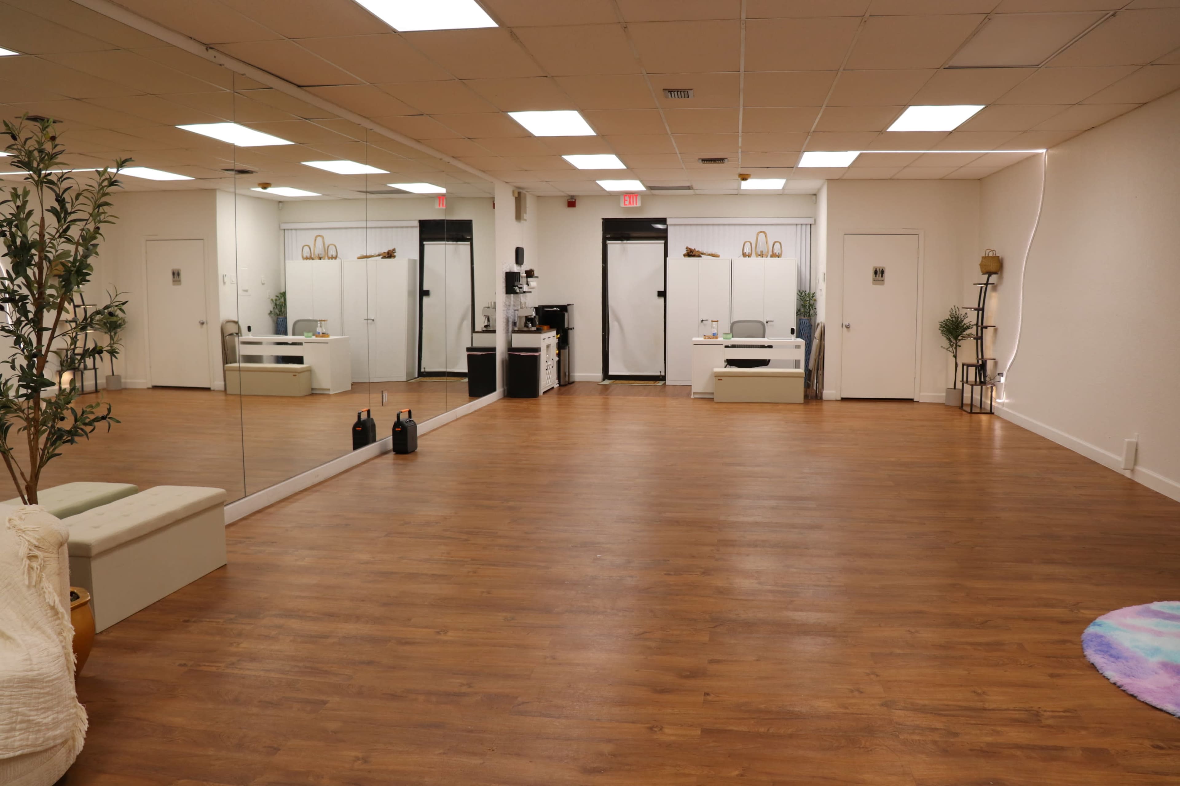 The image shows a spacious studio with a wooden floor, a large mirror on one wall, and minimalist furniture including a bench and small white tables.