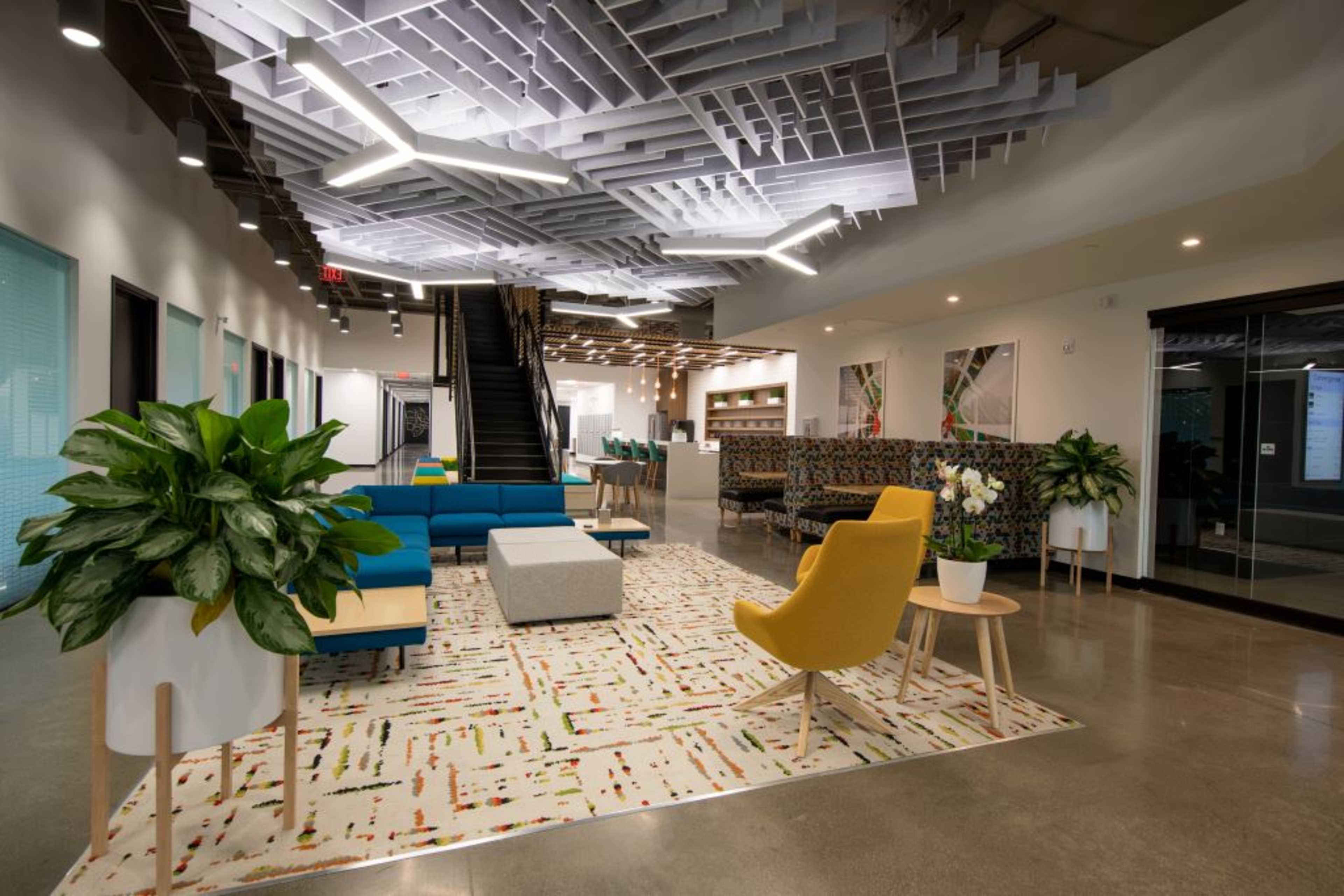 The image depicts a modern office lounge area featuring colorful seating, a large area rug, and a unique ceiling design.