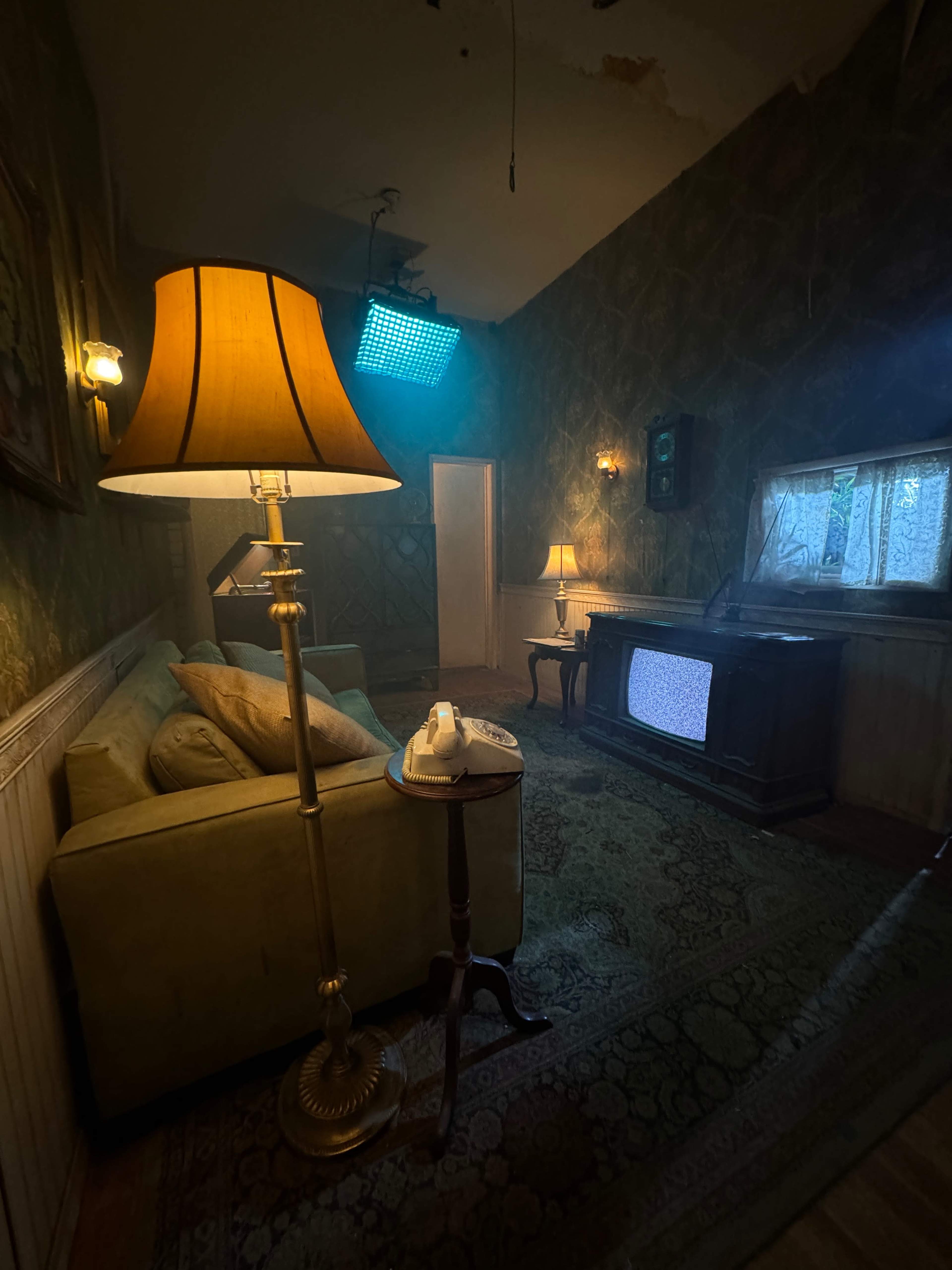 A dimly lit living room features a green sofa, an old television, a vintage rotary phone on a side table, and a standing lamp.