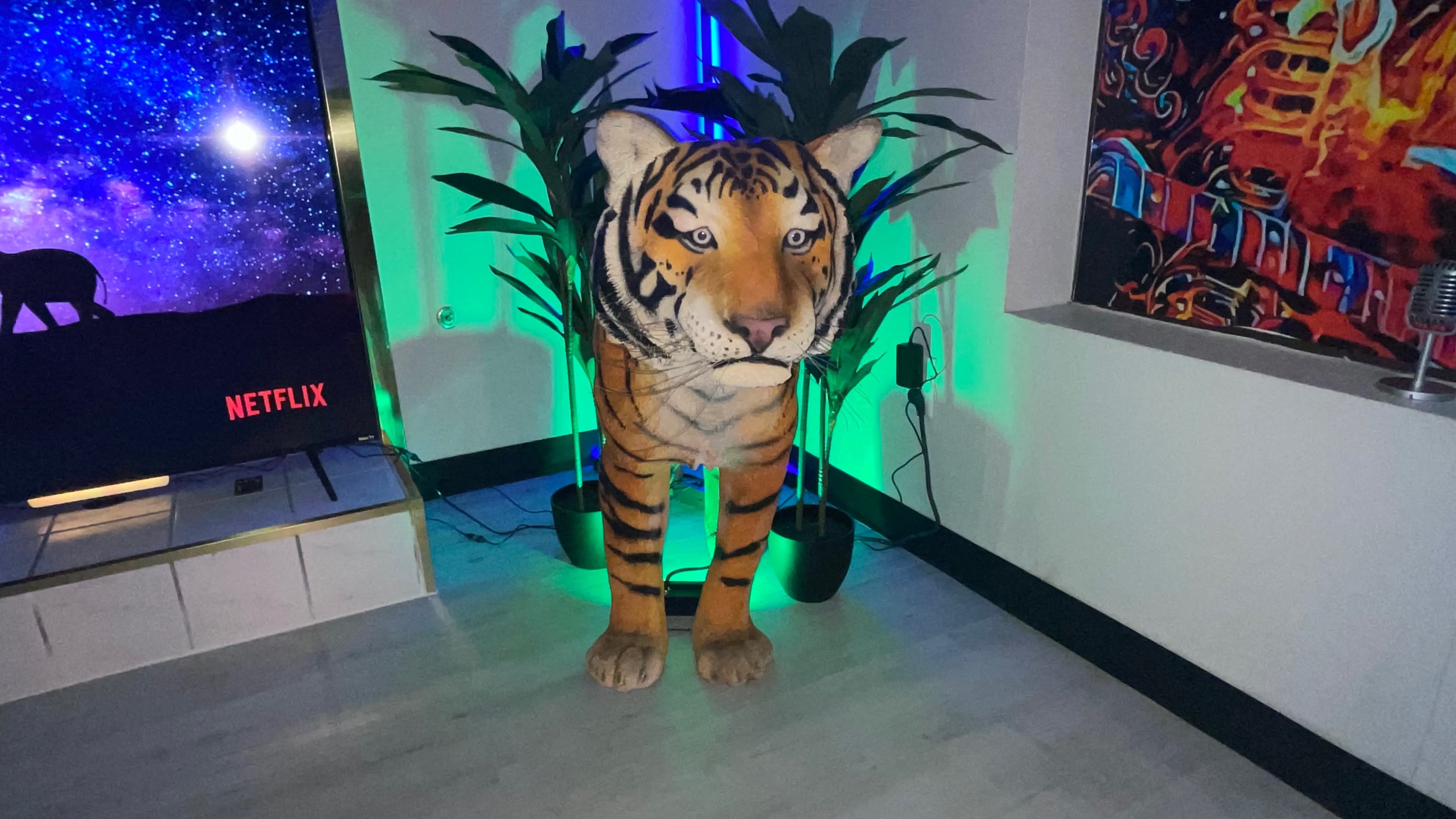 A life-sized tiger statue stands next to two plants, illuminated by colorful lights, in a room featuring a television displaying the Netflix logo.