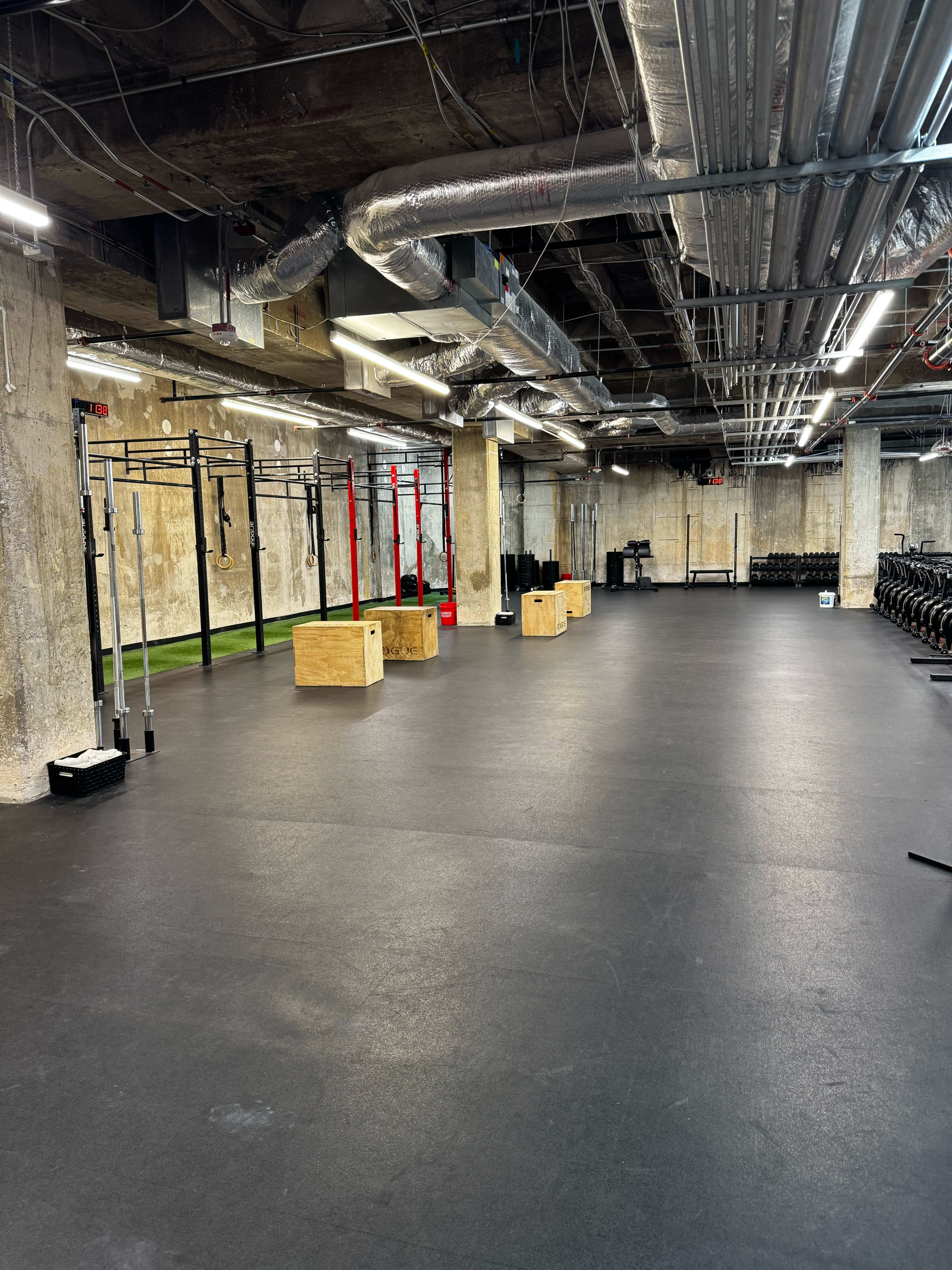 The image shows a spacious gym with various workout equipment, including box jumps and a pull-up rig, all set against a backdrop of exposed concrete walls and industrial-style ventilation.