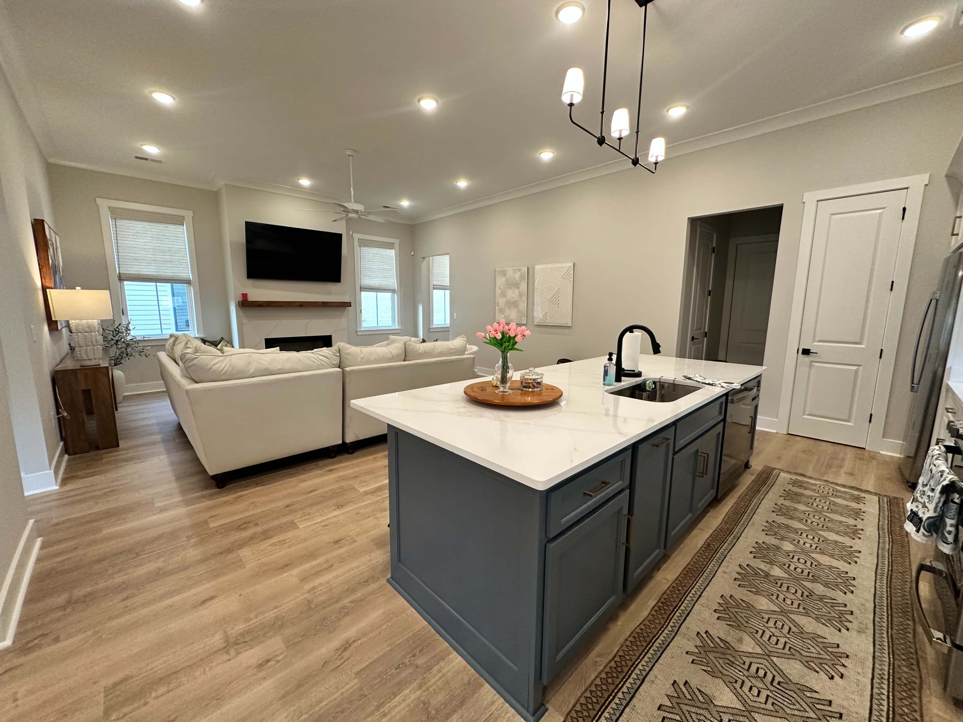 A modern kitchen features a central island with a marble countertop, facing a spacious living area with a light-colored sofa and a wall-mounted television.