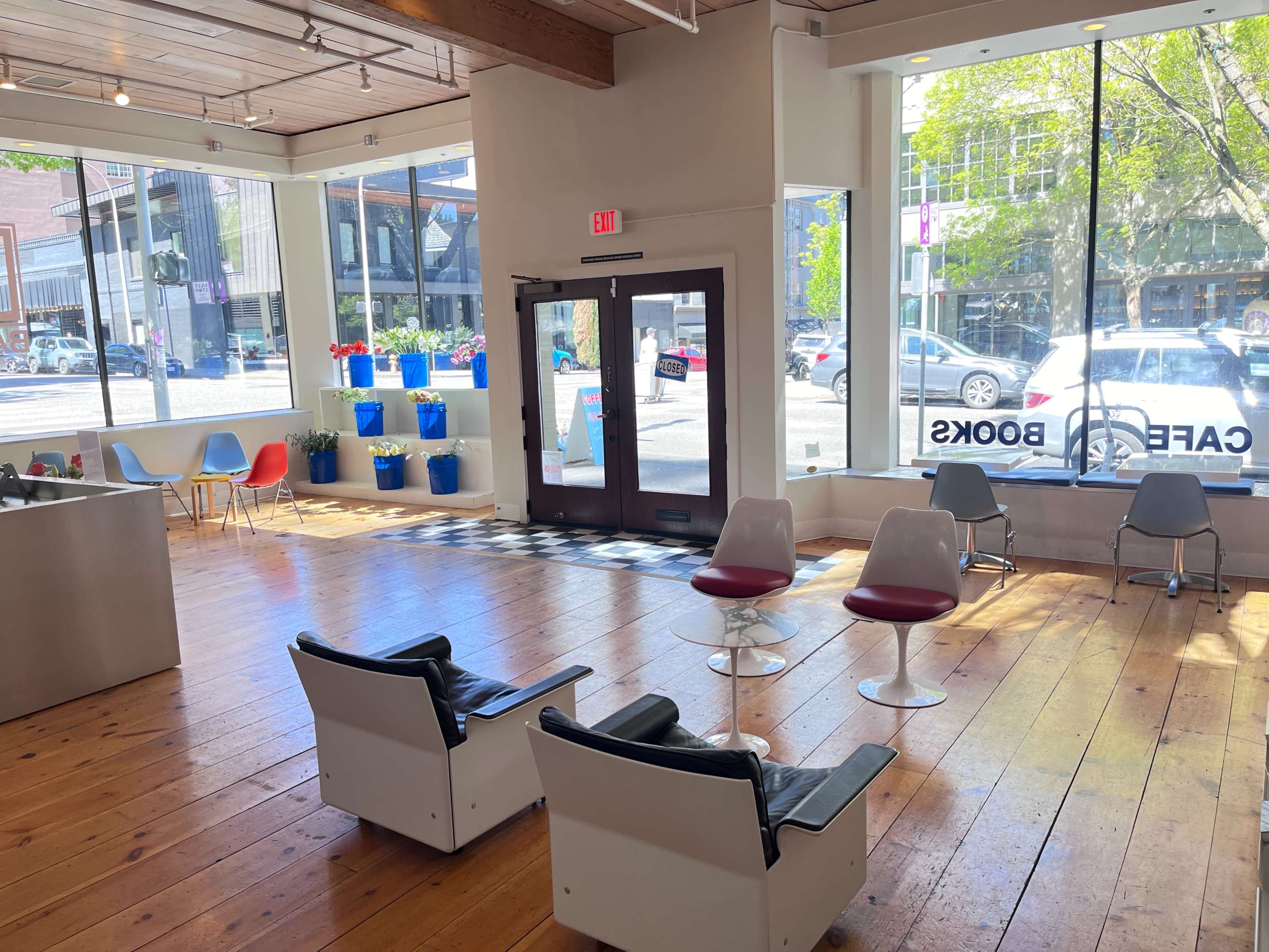 The image shows a brightly lit interior of a café with wooden floors and large windows, featuring modern seating and potted plants.