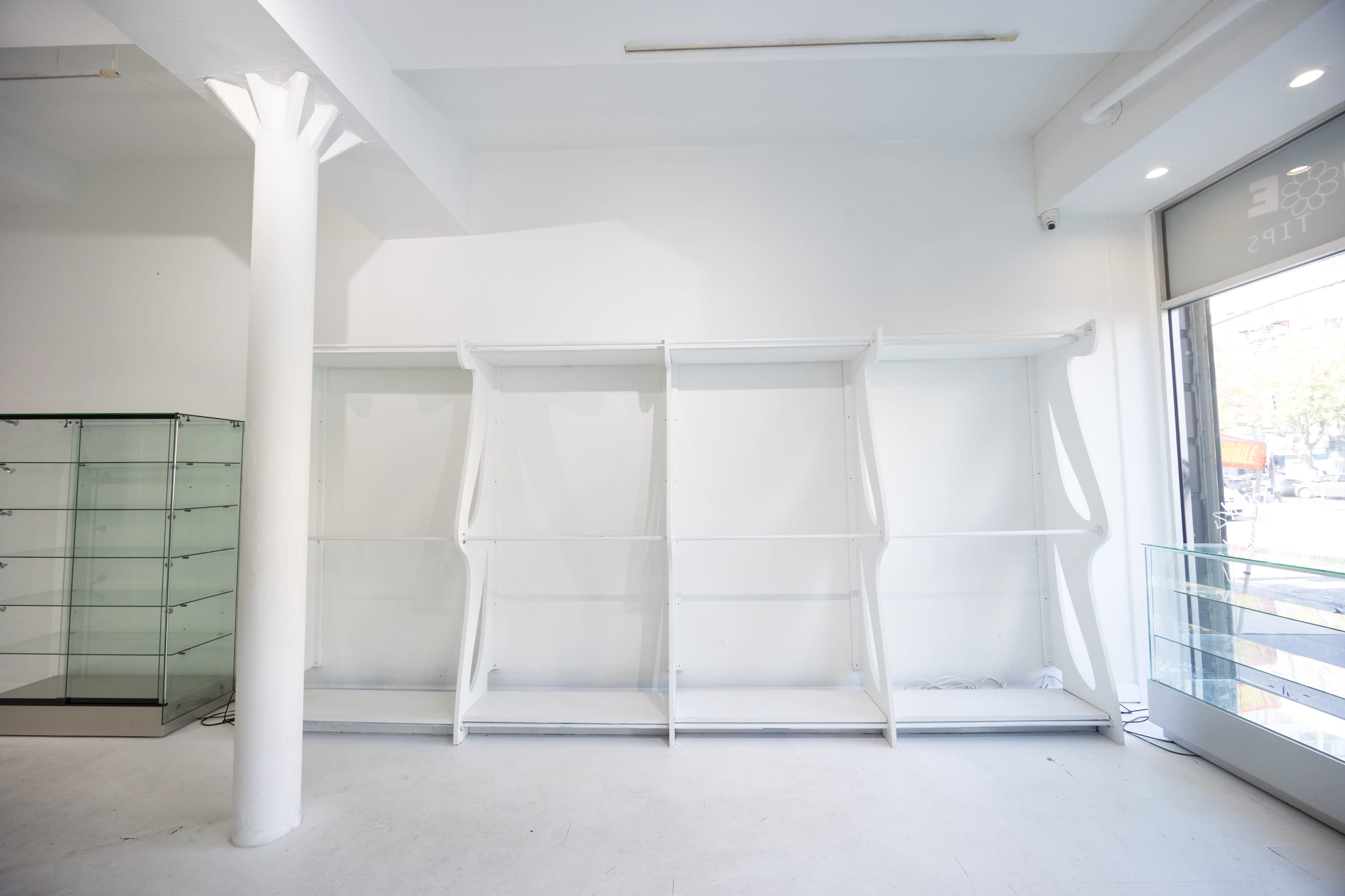 The image shows a minimalist interior of a store with empty white shelves against a plain white wall and a glass display section nearby.