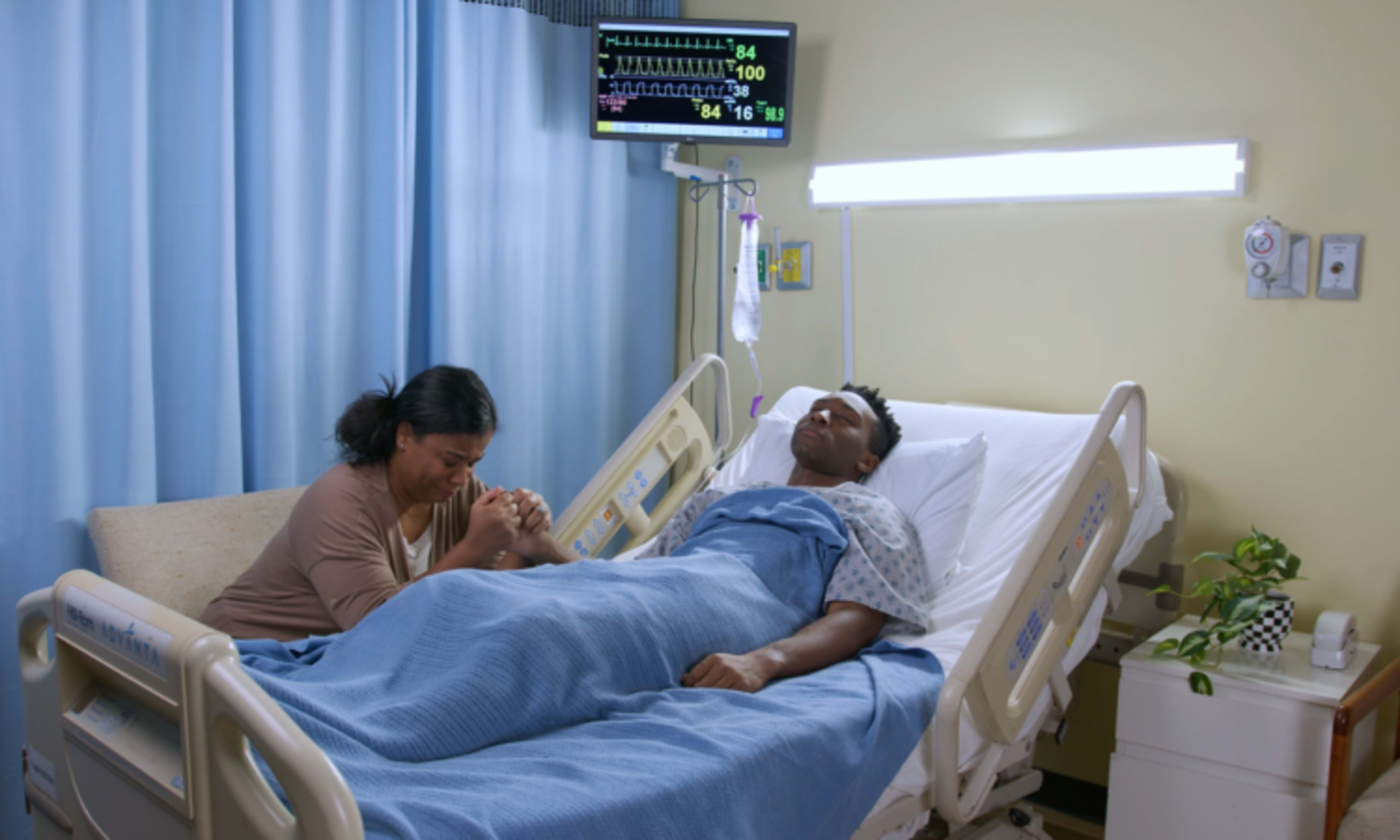 A woman sits beside a patient in a hospital bed, holding his feet while medical equipment monitors display readings.