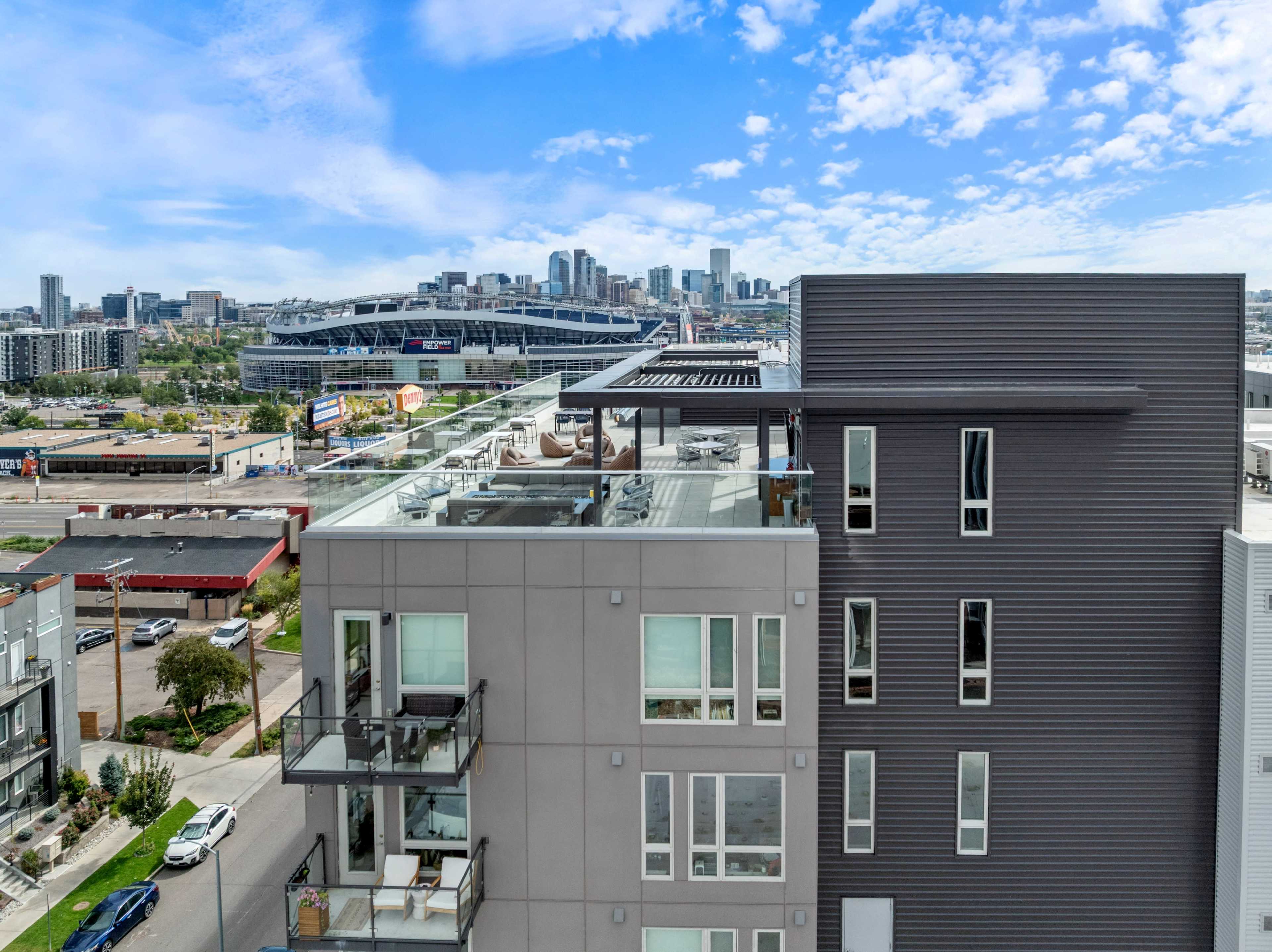 The image shows a modern multi-story building with a rooftop terrace overlooking a city skyline and stadium in the background under a partly cloudy sky.