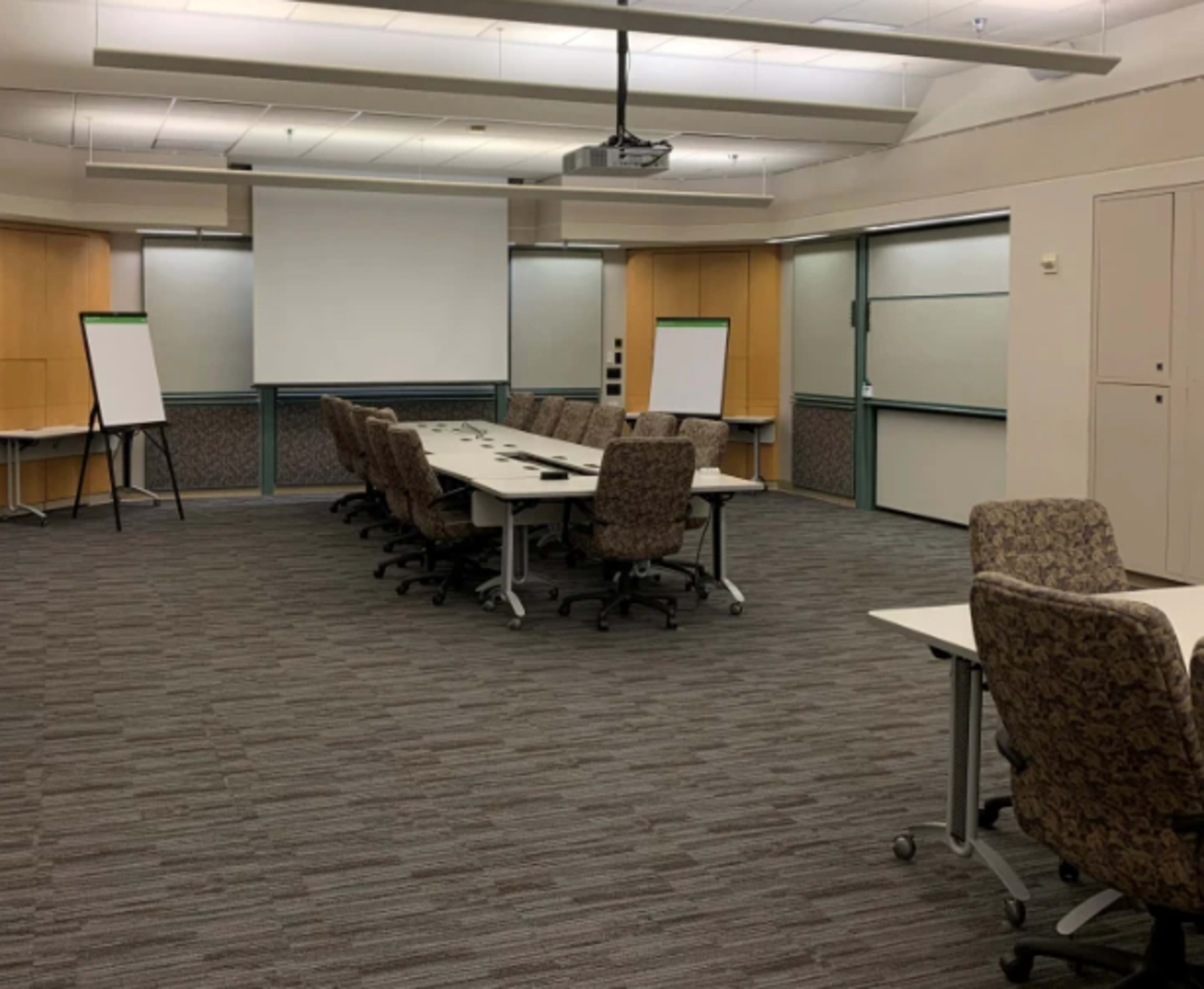 A conference room with a long meeting table, several office chairs, a projector, and empty whiteboards against the walls.