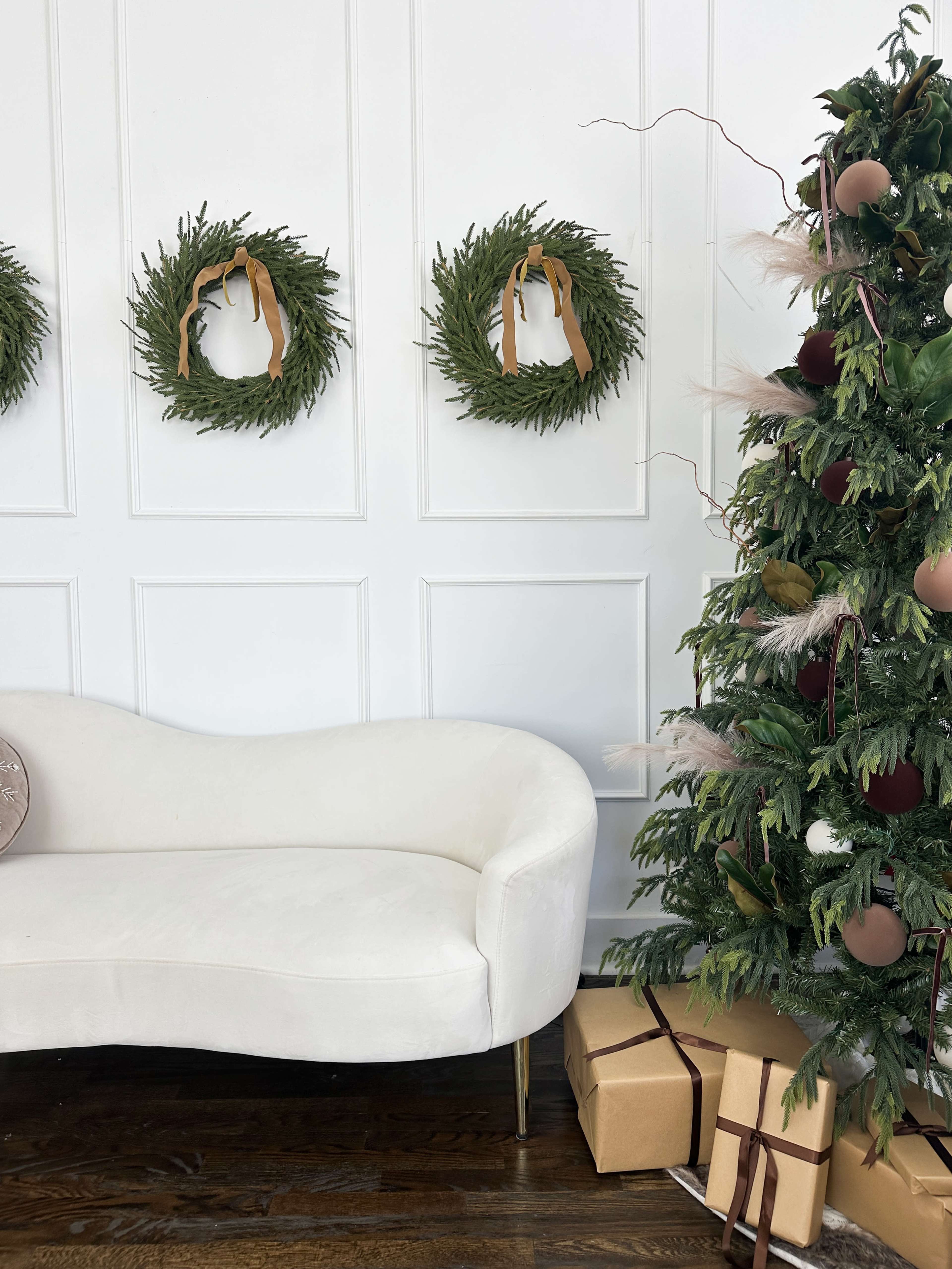A white curved sofa is positioned next to a decorated Christmas tree adorned with ornaments and surrounded by wrapped gifts, while three green wreaths hang on the white wall behind.