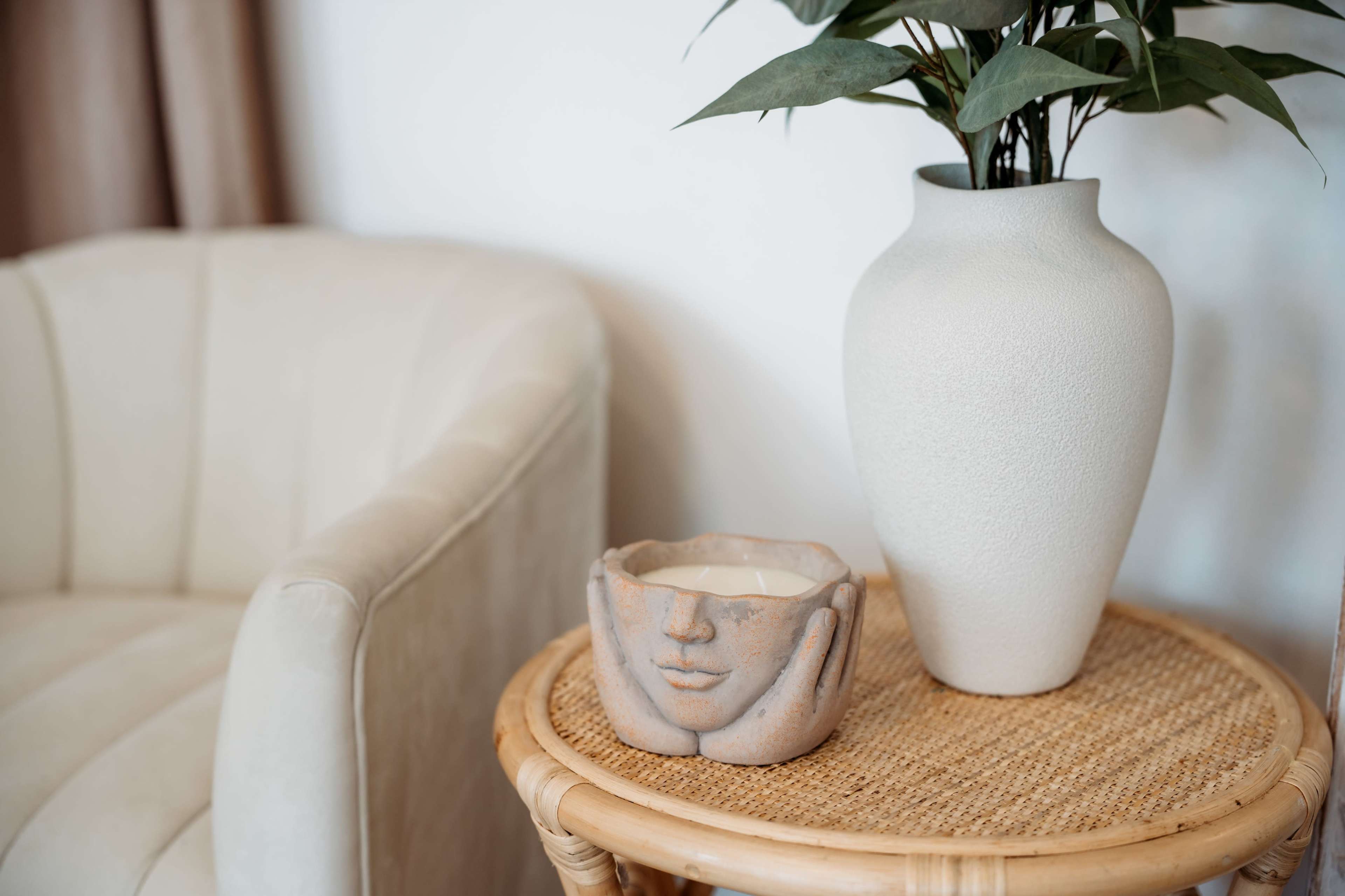 A cozy armchair sits beside a rattan side table topped with a face-shaped candle holder and a vase filled with greenery.