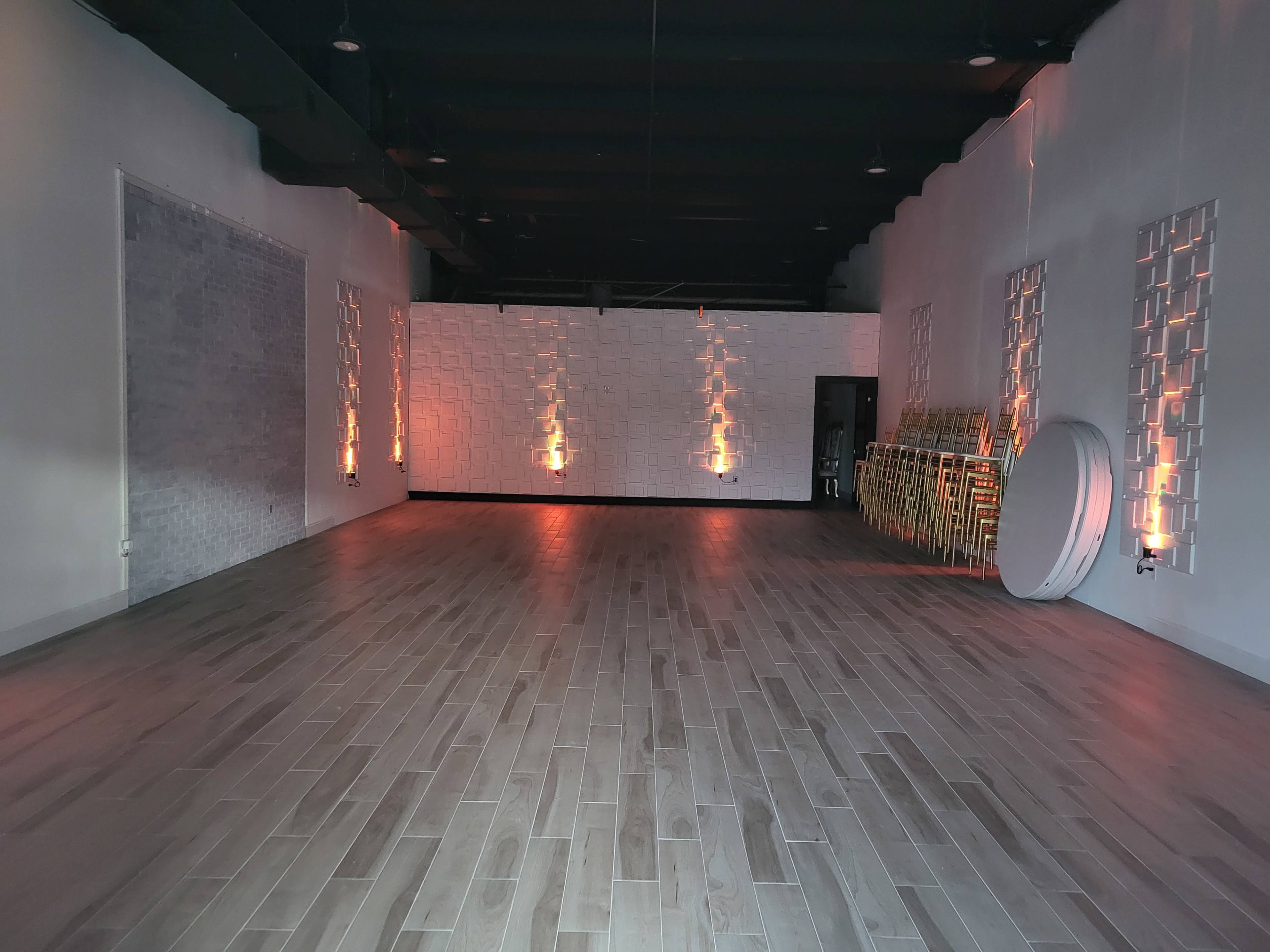 The image shows a spacious, empty room with wooden flooring, illuminated by soft orange lights along the walls and stacked chairs in the corner.