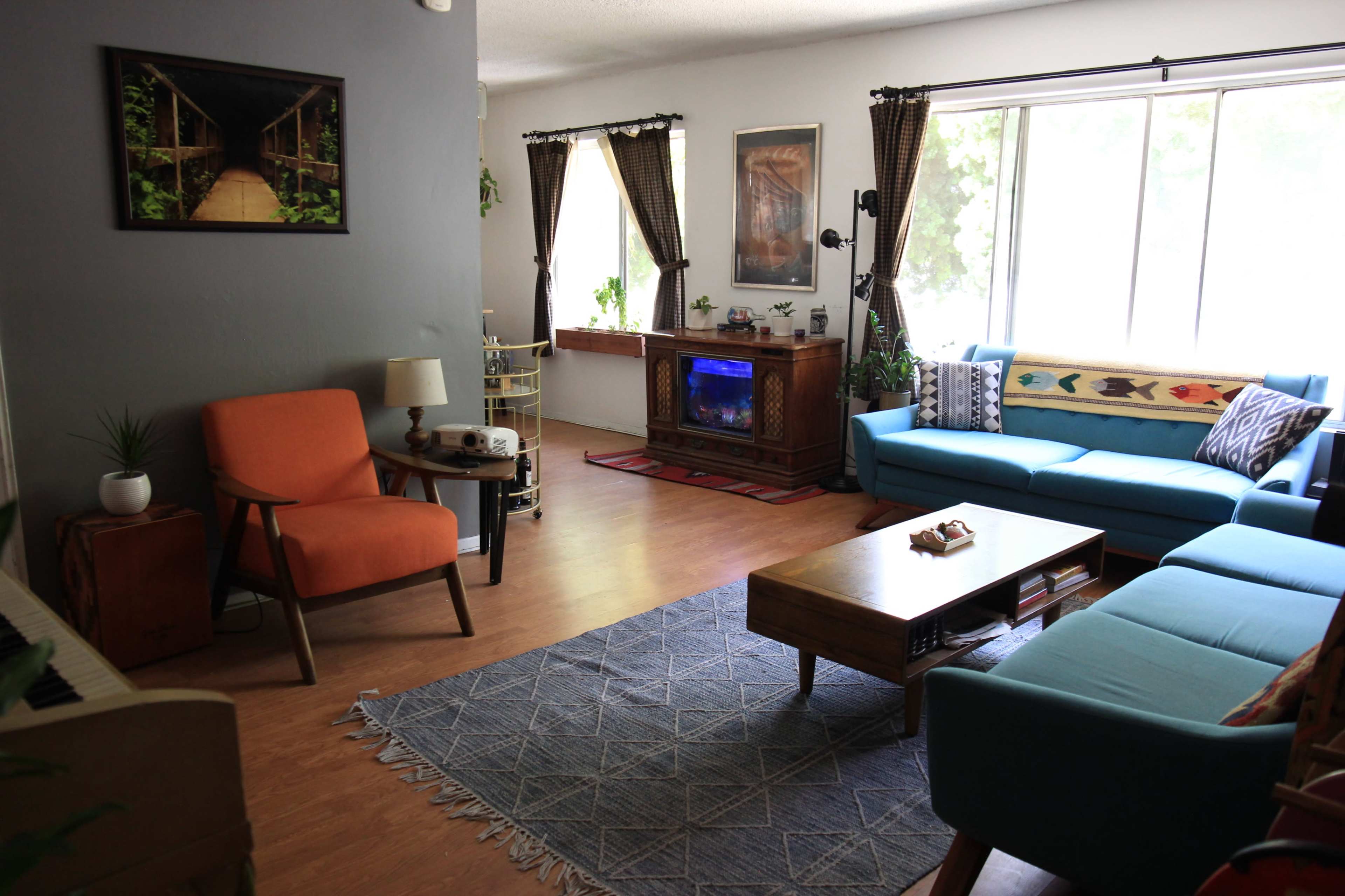 The living room features a blue sofa, an orange armchair, a wooden coffee table, and large windows that provide natural light.