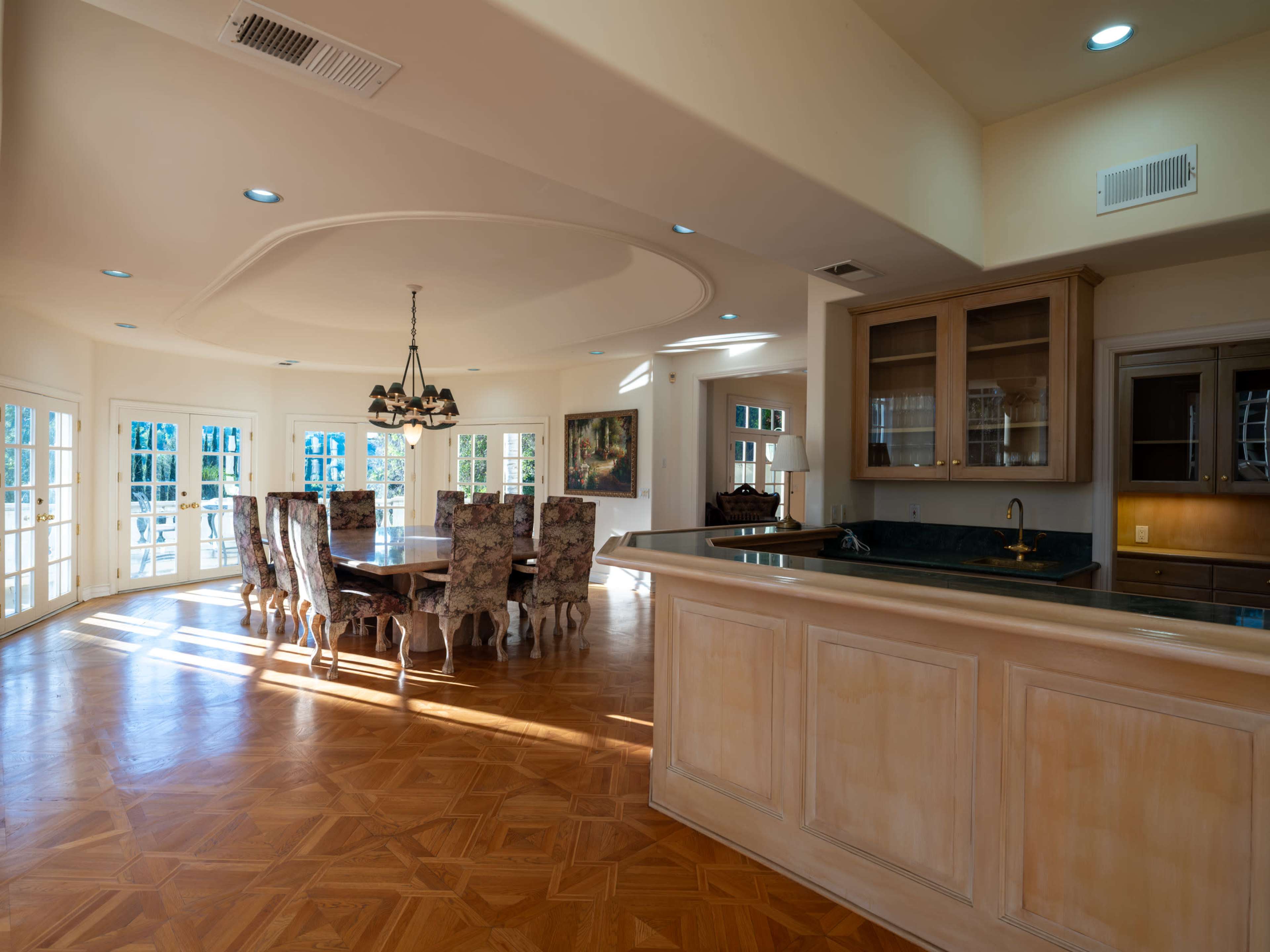 The image shows an open-concept dining area with a circular ceiling, a large wooden dining table surrounded by upholstered chairs, and a kitchen counter with cabinetry in the background.