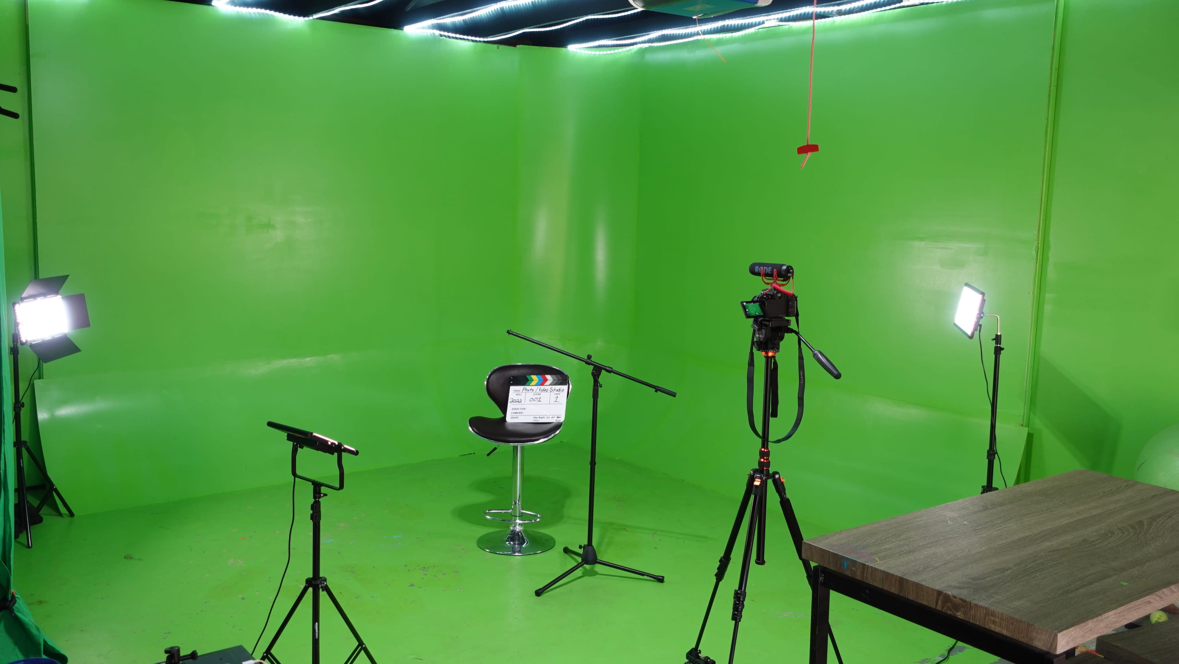 The image shows a green screen studio setup featuring a black chair, camera equipment, and lighting stands arranged around a central focus area.