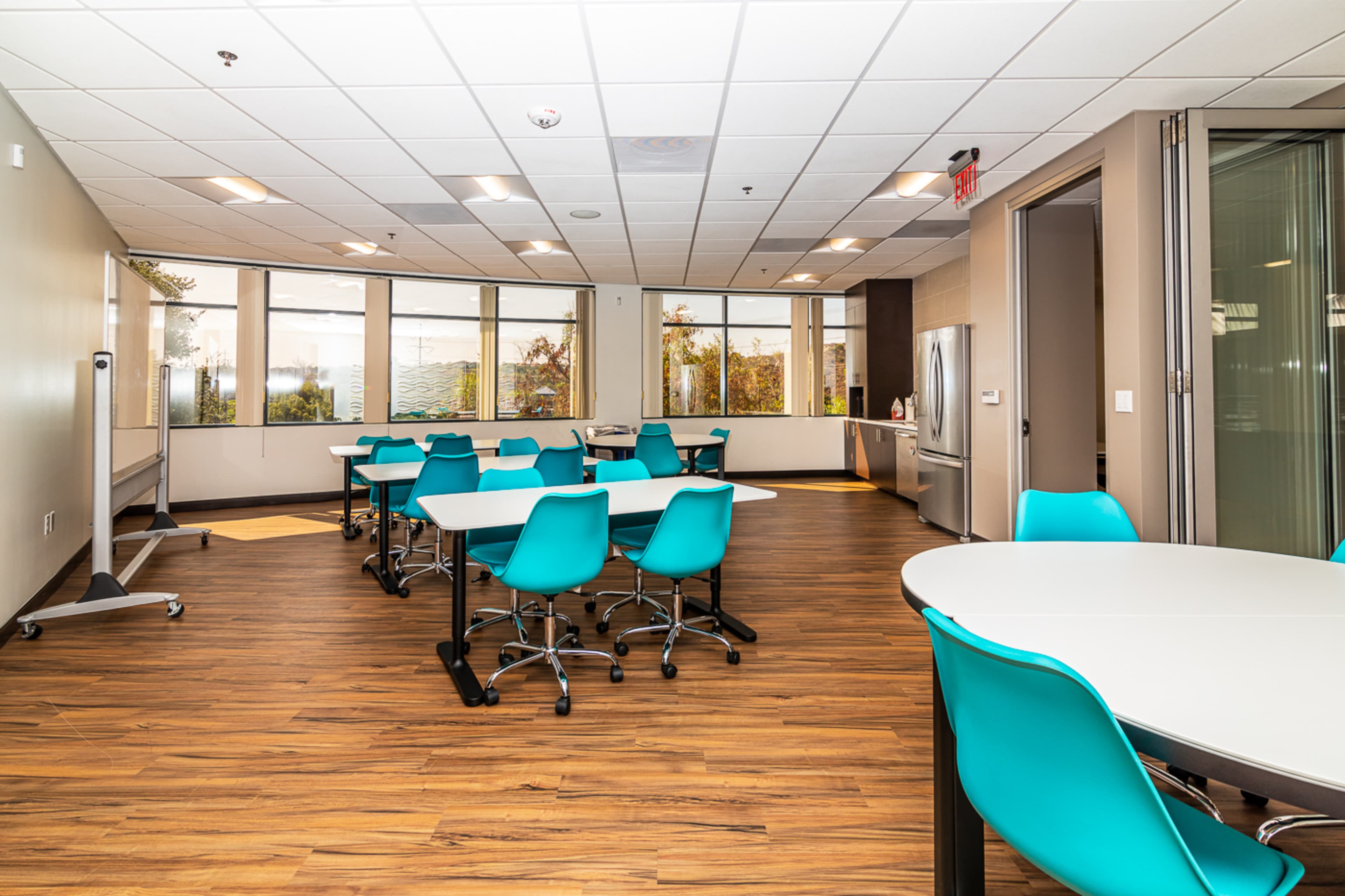 The image shows a brightly lit meeting room with several round tables and turquoise chairs, surrounded by large windows.