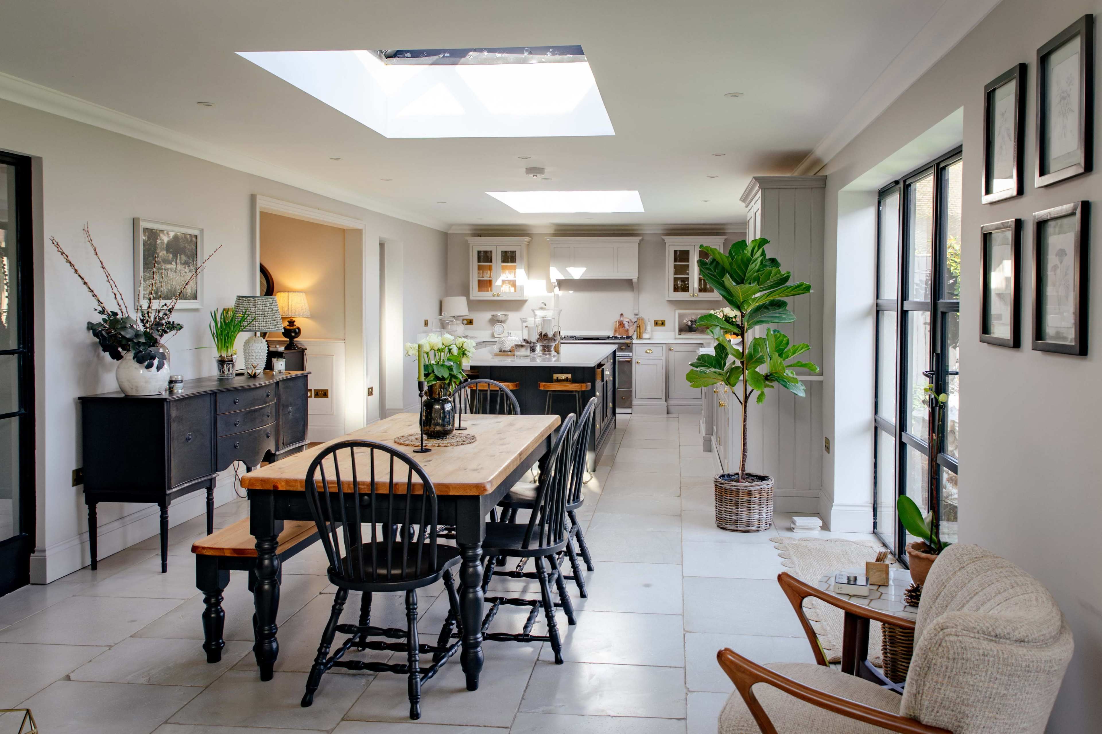 The image shows a modern kitchen and dining area featuring a large wooden table surrounded by black chairs, with a skylight above and large windows allowing natural light.