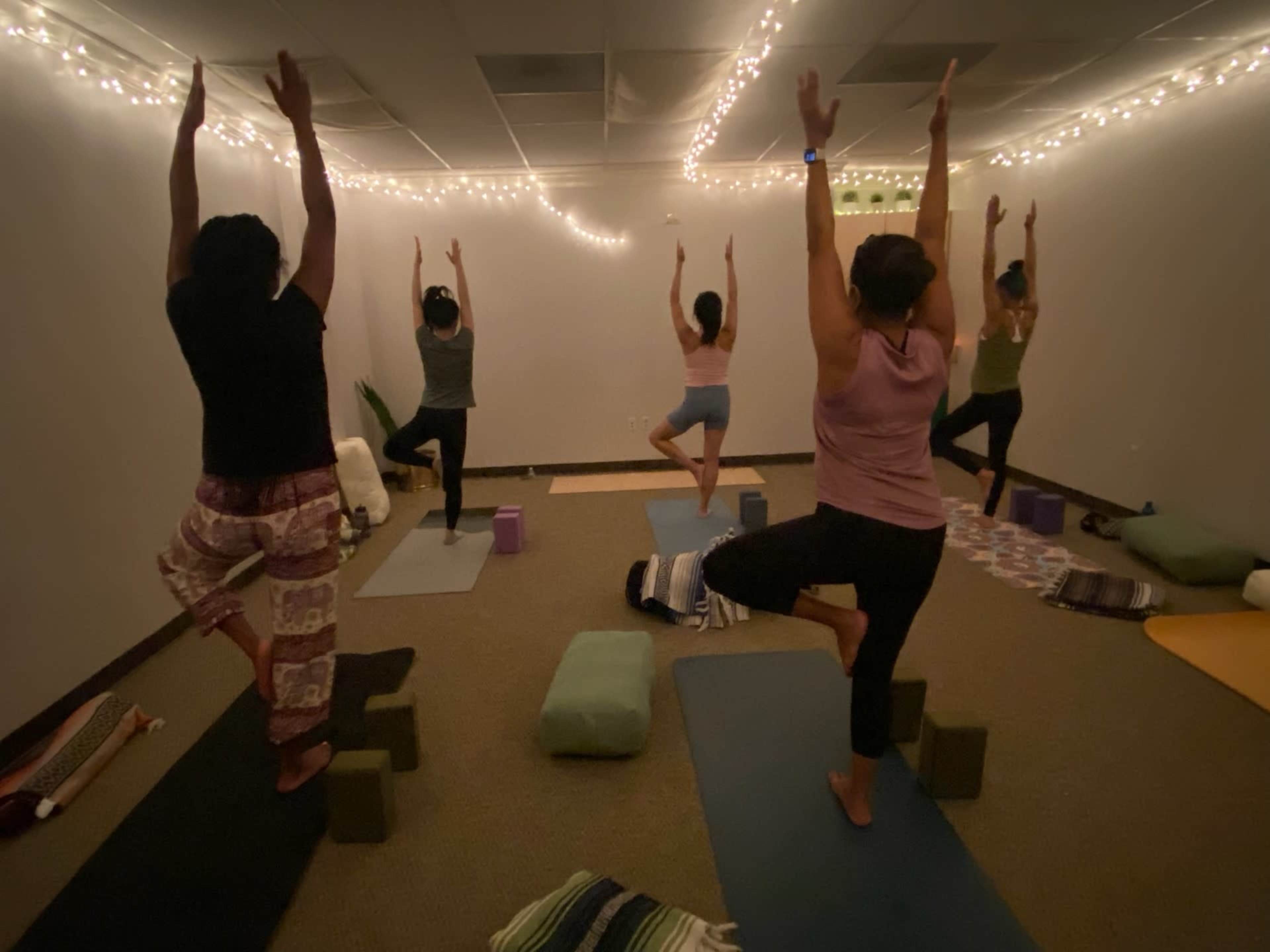 A group of five individuals performs yoga poses in a dimly lit studio adorned with string lights.