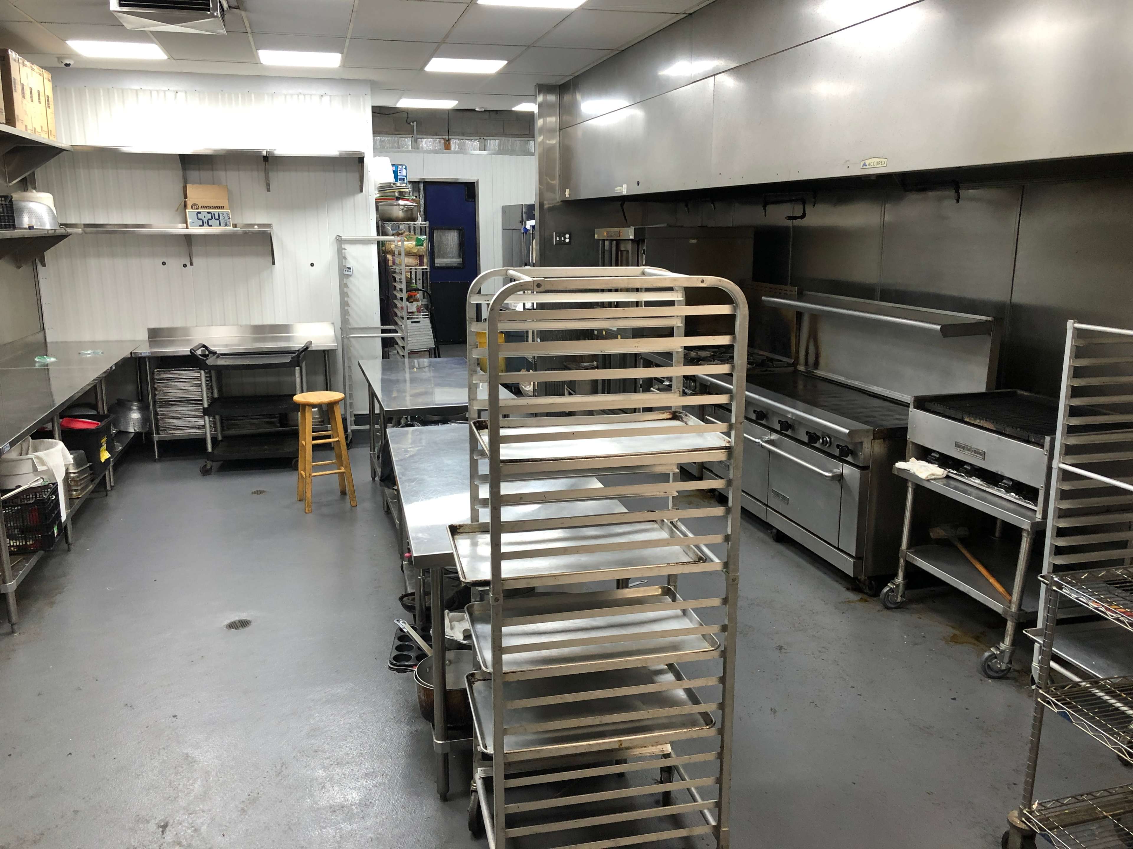 The image shows a commercial kitchen with stainless steel equipment, shelves, and a stool arranged in a spacious layout.