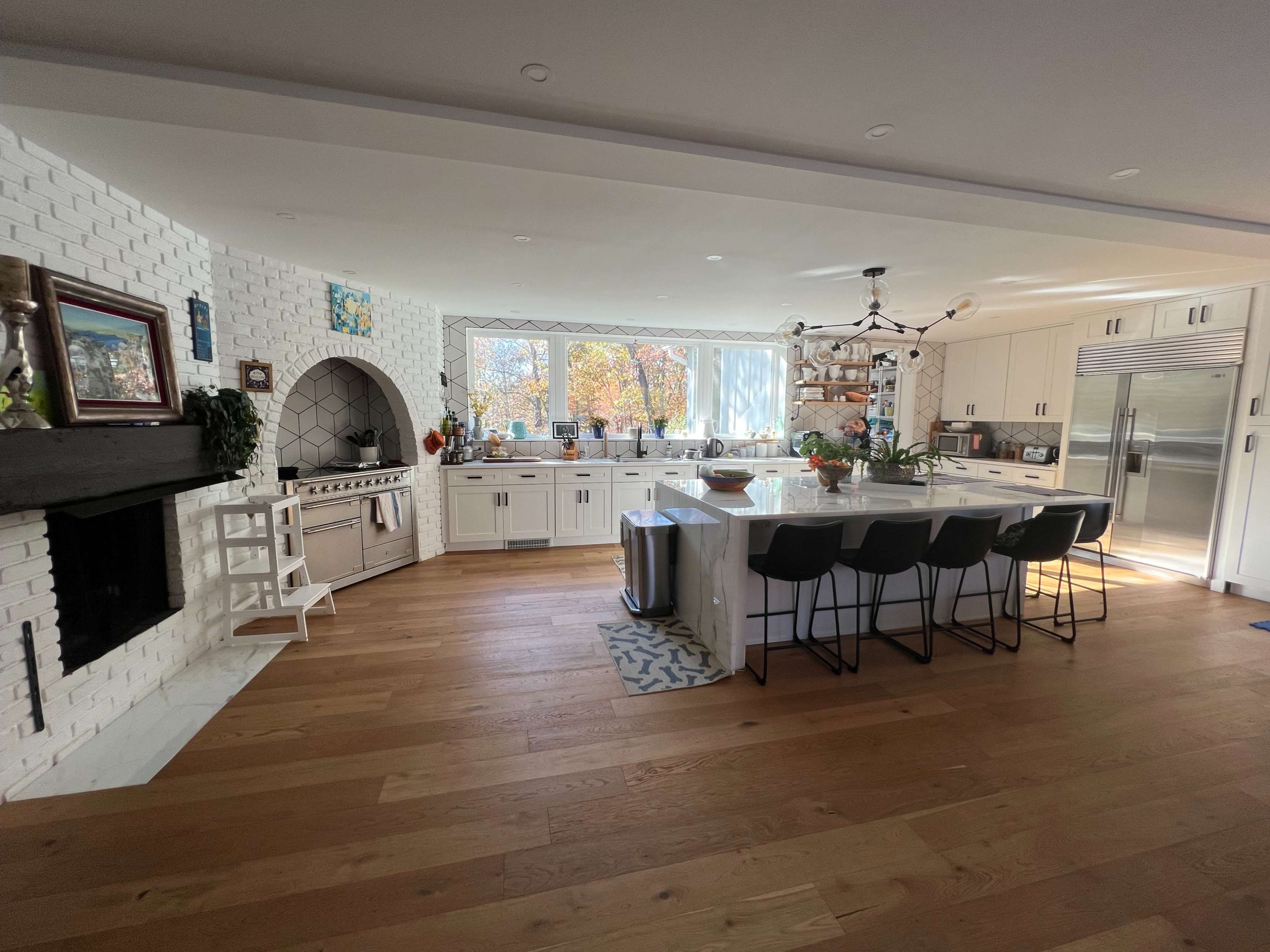 The image shows a spacious kitchen with white brick walls, a large island with black stools, and ample natural light coming through a wide window.