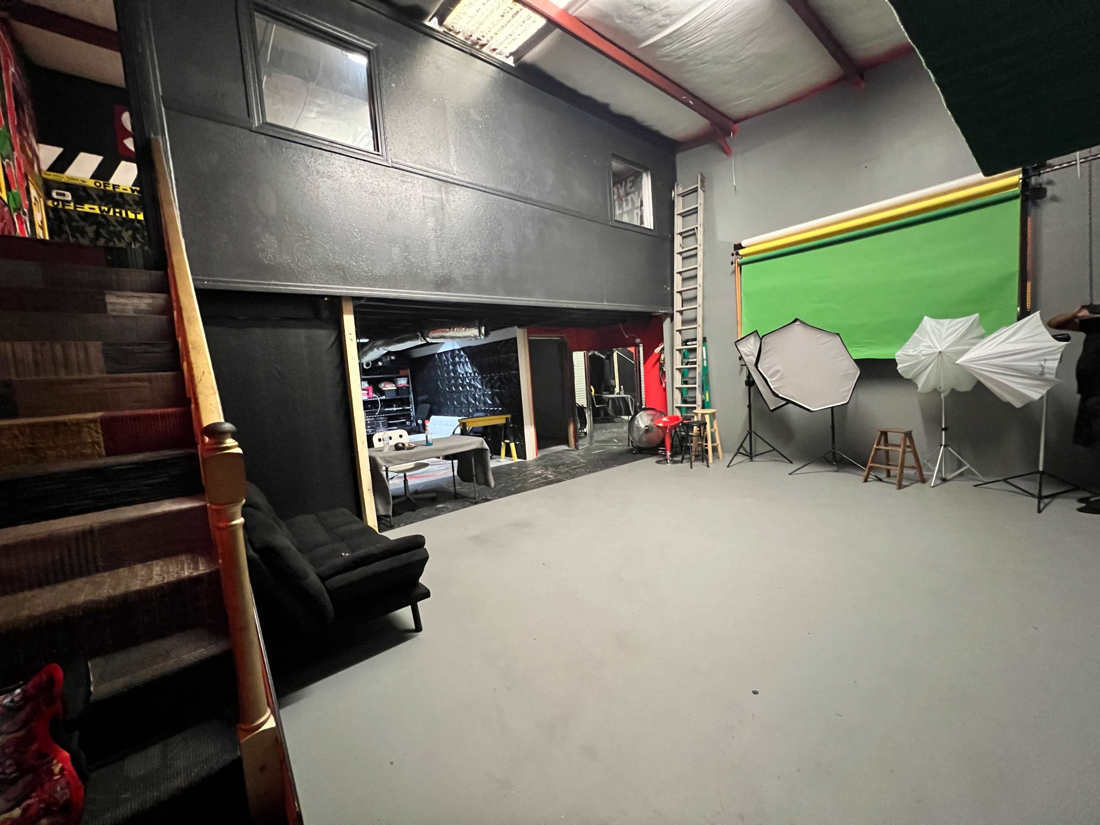 A spacious studio with a gray floor, black walls, a green backdrop, various lighting equipment, and a staircase leading to an upper level.