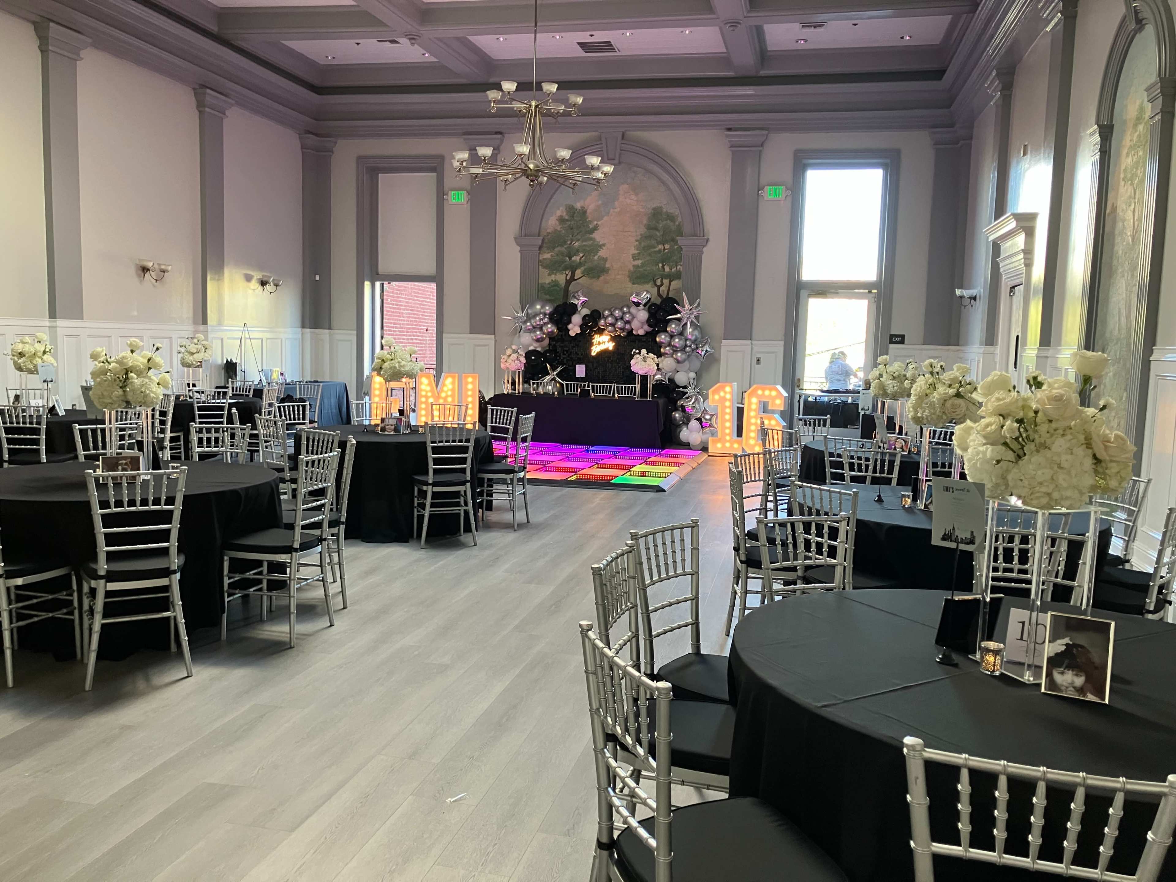 The image shows a banquet hall set up with round tables covered in black tablecloths, silver chiavari chairs, and a stage area decorated with flowers and illuminated letters spelling "PM."