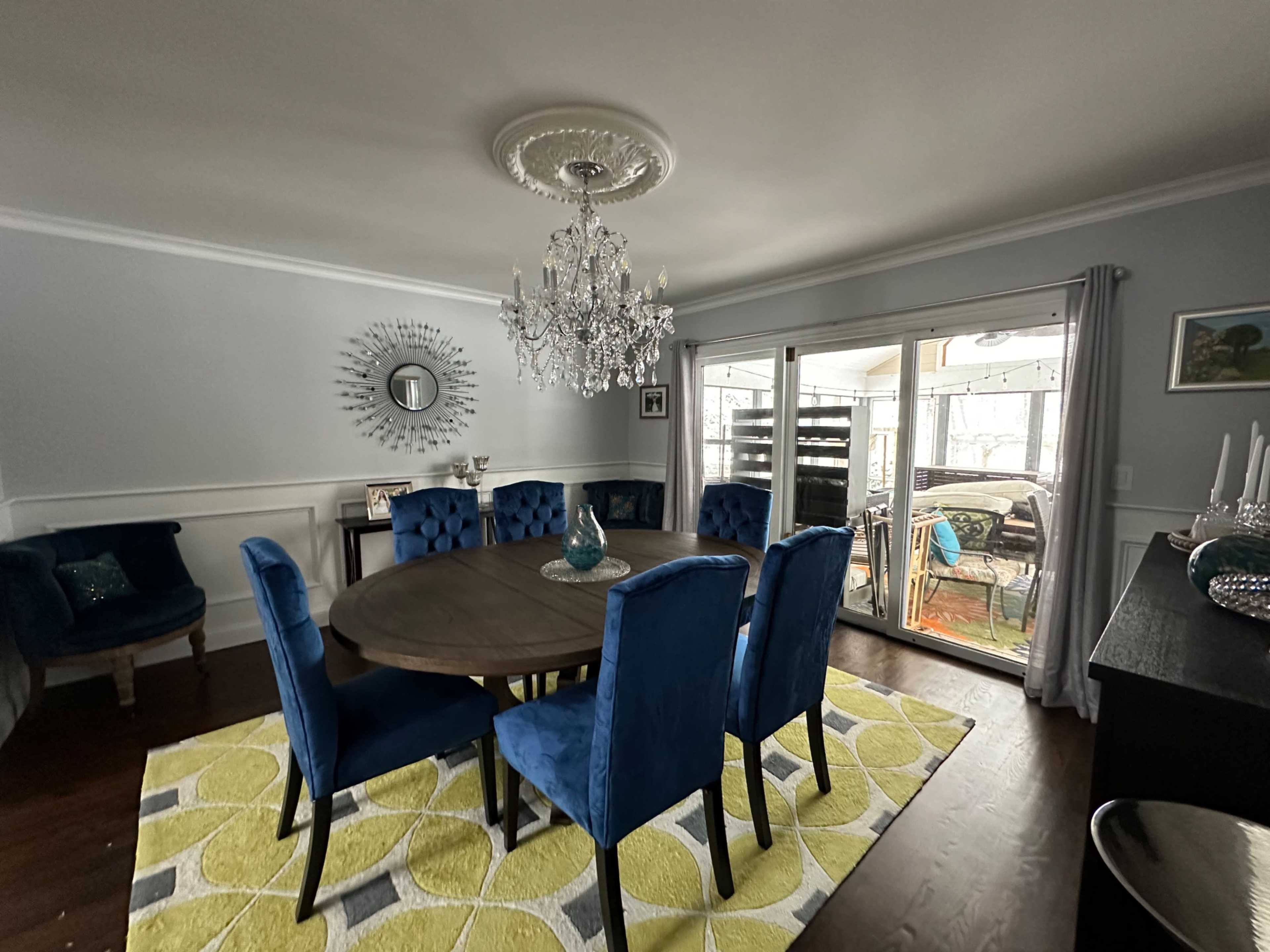 A dining room features a round wooden table surrounded by blue velvet chairs, a chandelier above, and sliding glass doors leading to an outdoor space.