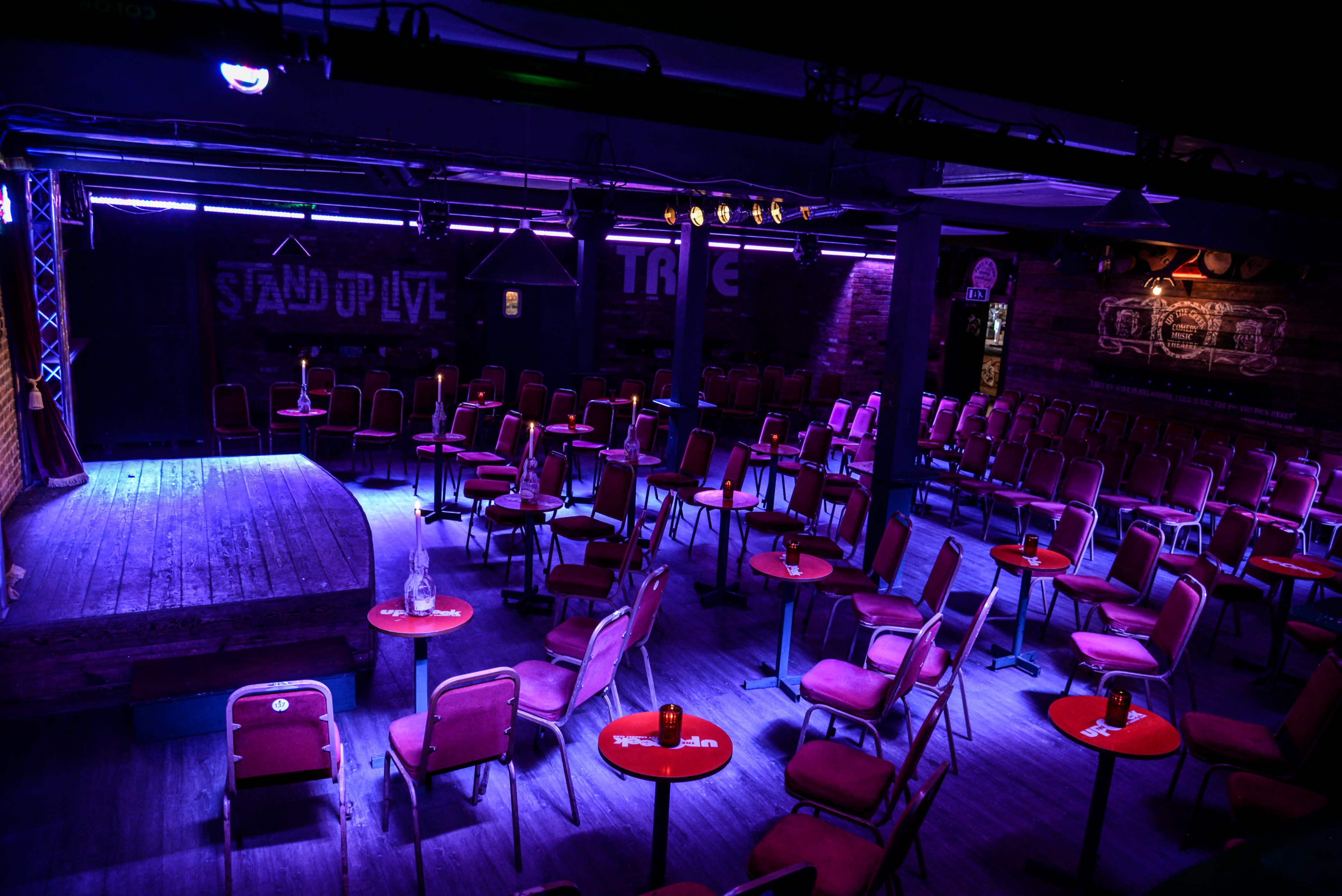 The image shows a dimly lit comedy venue with rows of empty chairs and small round tables facing a small stage.