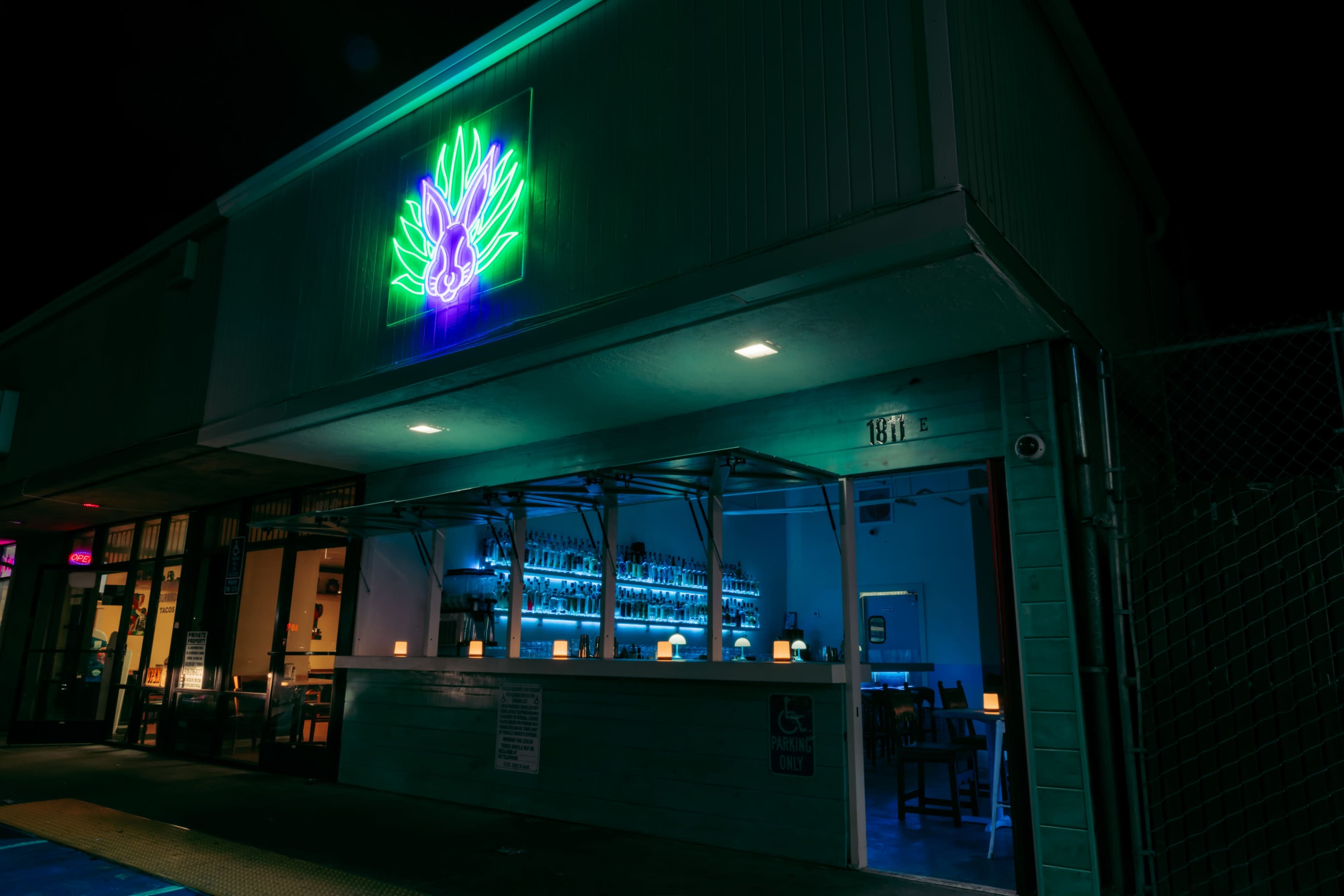 A brightly lit bar exterior features a neon pineapple sign and illuminated shelves filled with bottles inside.