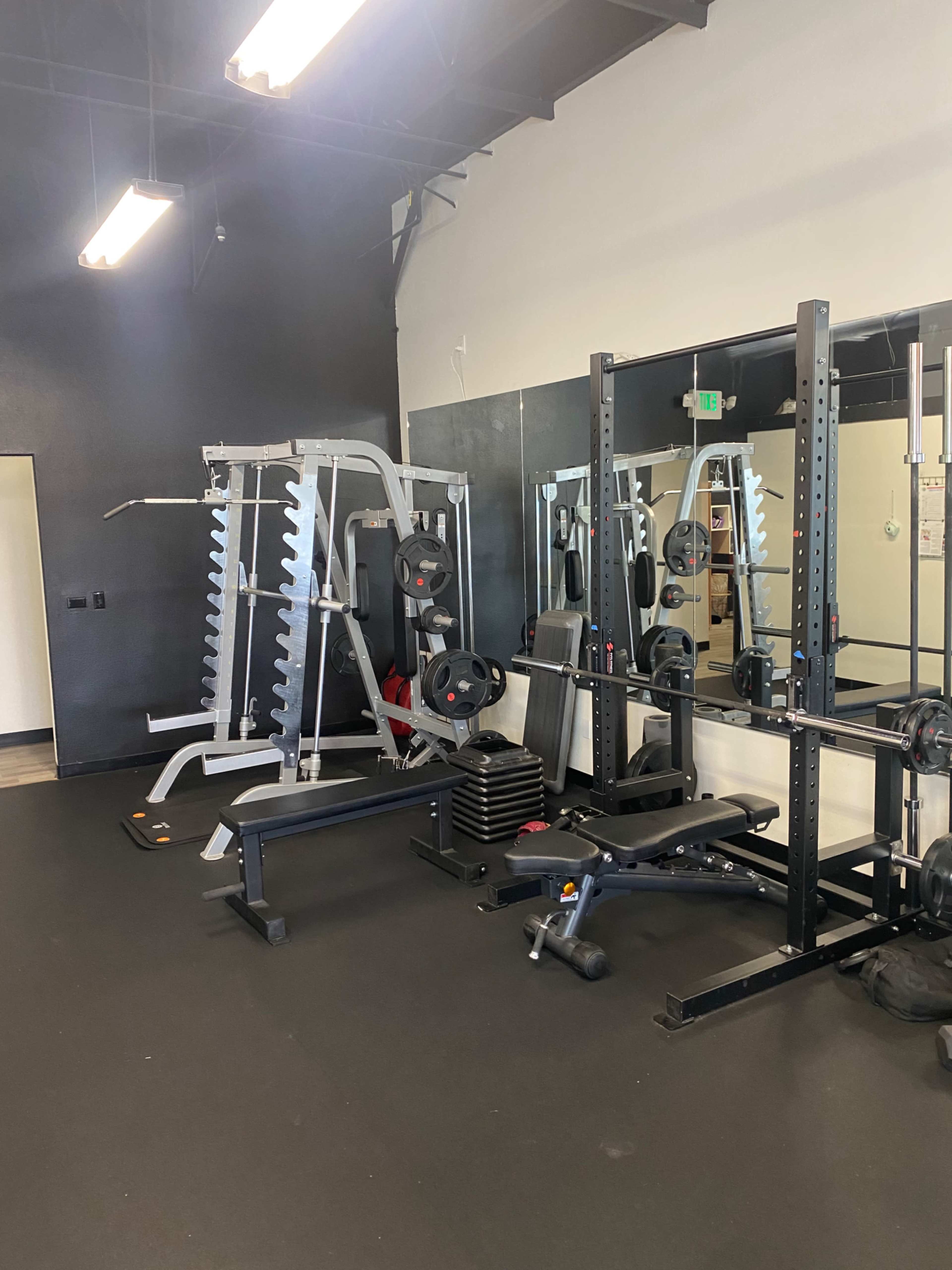 A gym interior with various weightlifting equipment, including a squat rack, bench press, and weights, against a mirrored wall.